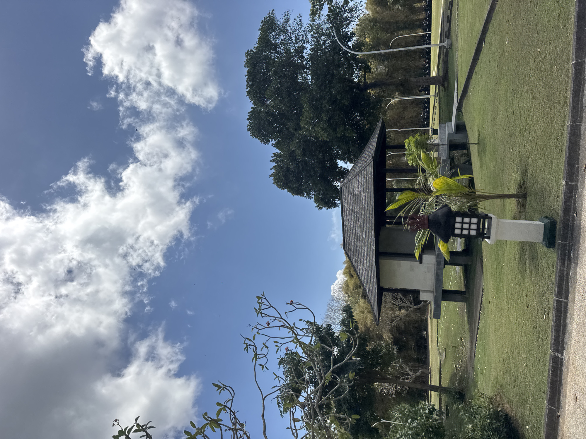 The image captures an outdoor scene in Kuta Selatan, Indonesia, under a bright blue sky with scattered white clouds. In the foreground, a well-maintained lawn surrounds a small, elevated, open-sided pavilion with a dark, shingled roof. A decorative lantern sits on a white pedestal to the left of the pavilion. The pavilion is supported by dark pillars, and a small bridge leads to it from the right. Beyond the pavilion, a large, dense green tree dominates the middle ground, with other trees and vegetation forming a backdrop. In the far background, a line of motorcycles is visible, suggesting proximity to a road or a parking area. The overall impression is one of a serene, park-like environment.