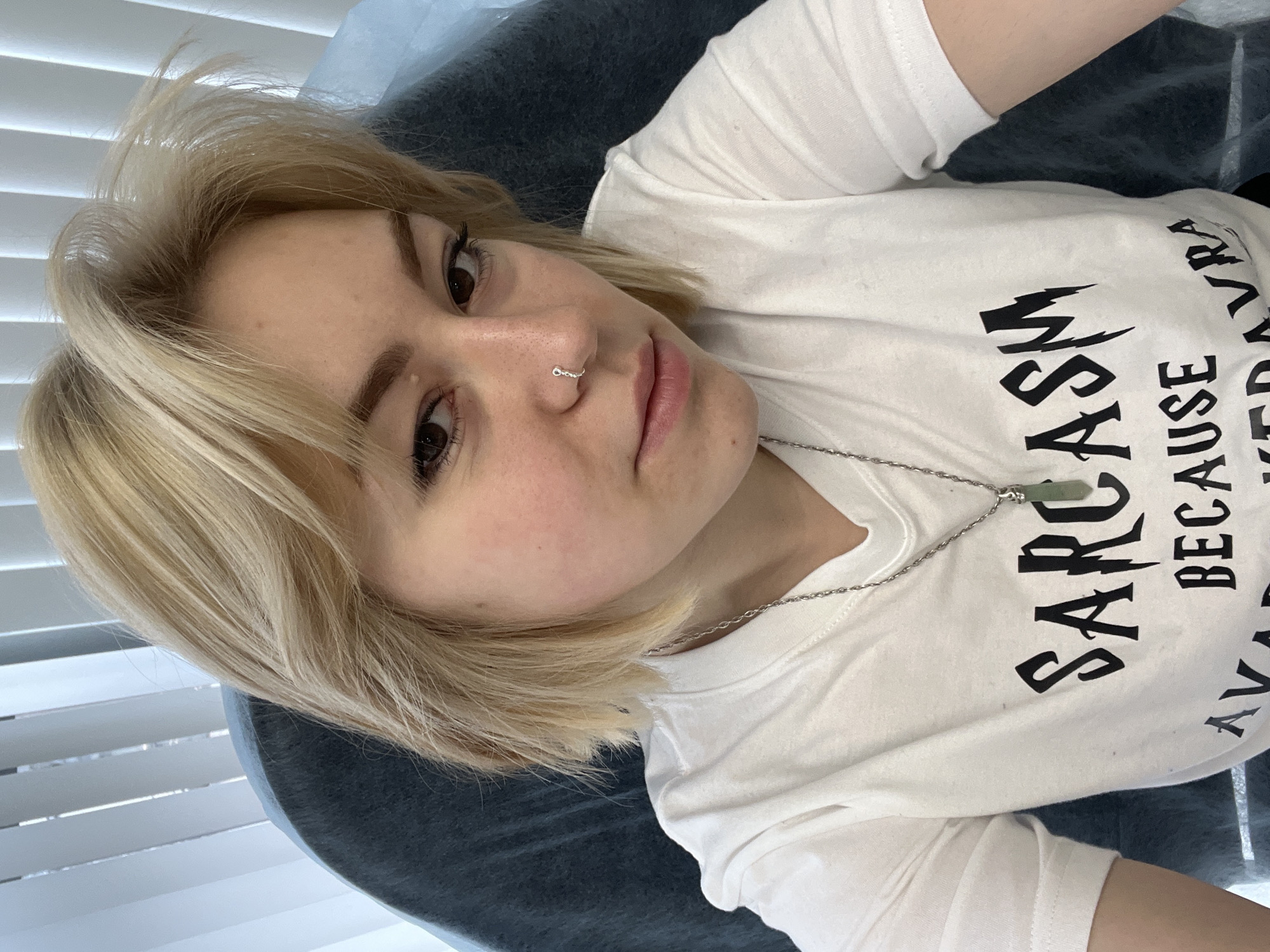 A young woman with short, light blonde hair and brown eyes is captured in a selfie taken from an overhead perspective, looking directly at the camera with a neutral, relaxed expression. She has fair skin, faint freckles across her nose and cheeks, and a small silver ring piercing on her right nostril.
She is wearing a plain white t-shirt adorned with black, stylized lettering. The most prominent word visible is "SARCASM," followed by "BECAUSE" and parts of additional text that appear to be "AVADA KEDAVRA," suggesting a humorous slogan often associated with the Harry Potter series. Around her neck is a silver chain necklace from which a rectangular, light green pendant, possibly jade or aventurine, hangs.
The woman is lying on a dark grey or blue padded surface, which could be a bed, couch, or examination table. To her left, white horizontal blinds are visible in the background, indicating an indoor setting with natural light, likely daytime. The image provides no specific visual indicators of the location Odesa, Ukraine, beyond the subject herself.