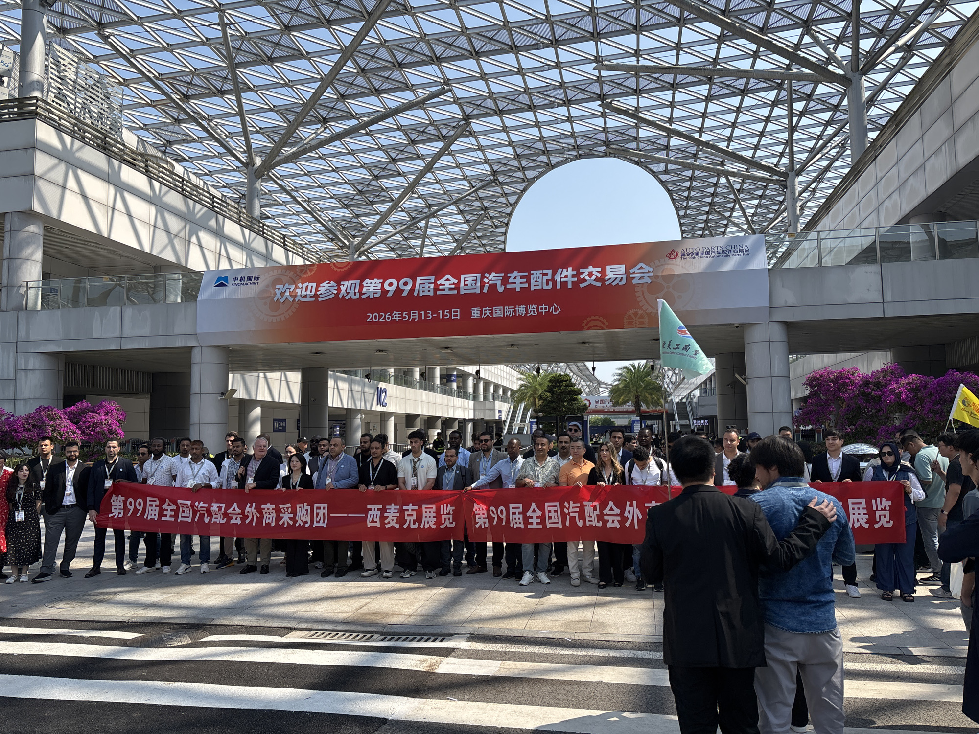 A group of people are holding a long red banner that reads "第99届全国汽配会外商采购团————西麦克展览 第99届全国汽配会外" in Chinese characters. Above them, a larger red banner announces the 99th National Auto Parts Fair (全国汽车配件交易会), scheduled for May 13-15, 2026, at the Chongqing International Expo Center (重庆国际博览中心). The characters "中机国际 SINOMACHINT" also appear on the banner.
The scene is taking place in an urban environment with modern architecture. The building overhead has a large, complex glass and steel roof structure. There are trees and other greenery in the background. The event seems to be taking place in Liangjiang, China.