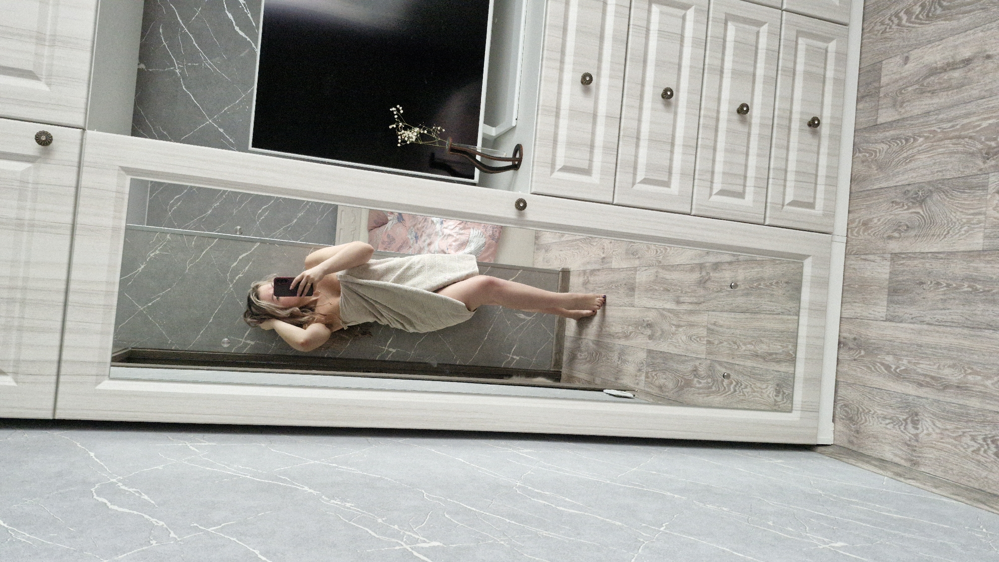 The image displays an indoor scene, likely a bedroom or dressing area in Otchor, Russia, captured as a mirror selfie. A person, appearing female, is reflected in a long, horizontal mirror integrated into a light-colored wooden cabinet or wardrobe unit. She is wrapped in a light-colored towel, covering her torso and part of her legs, with her face obscured by a dark-colored smartphone held up to take the picture. Her light brown hair falls around her shoulders, and her left leg is visible from the knee down, revealing a foot with dark-painted toenails.
The mirror reflects a background of dark grey wall tiles with prominent white, marble-like veining, and a glimpse of pink patterned fabric, possibly a bedspread or curtain. Above the mirrored section, the cabinet unit continues with matching light wood-paneled doors, each adorned with a circular, antique-style knob. A large black flat-screen television is mounted above the left portion of the cabinets, and to its right, a small wall-mounted vase holds delicate white flowers.
The floor in the immediate foreground consists of light grey tiles with white veining, similar to the reflected wall pattern. To the right of the cabinet unit, the flooring transitions to light-toned wooden laminate planks. The environment suggests a modern and tidy interior. No visible text is present in the image.