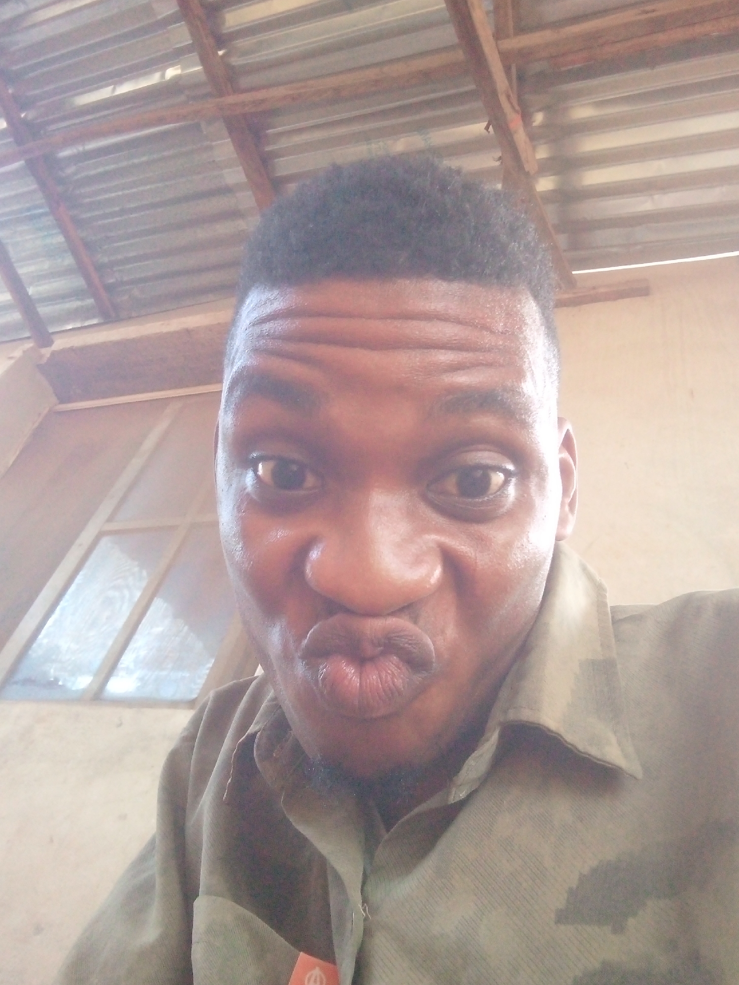 A man is seen in a selfie-style photograph. He has short, dark hair and dark eyes. His facial expression involves pursed lips. He is wearing a button-down shirt with a camouflage pattern. The background shows a corrugated metal roof and a window frame. The location appears to be an indoor space, possibly a building with a simple construction. The city is Unknown, Unknown.