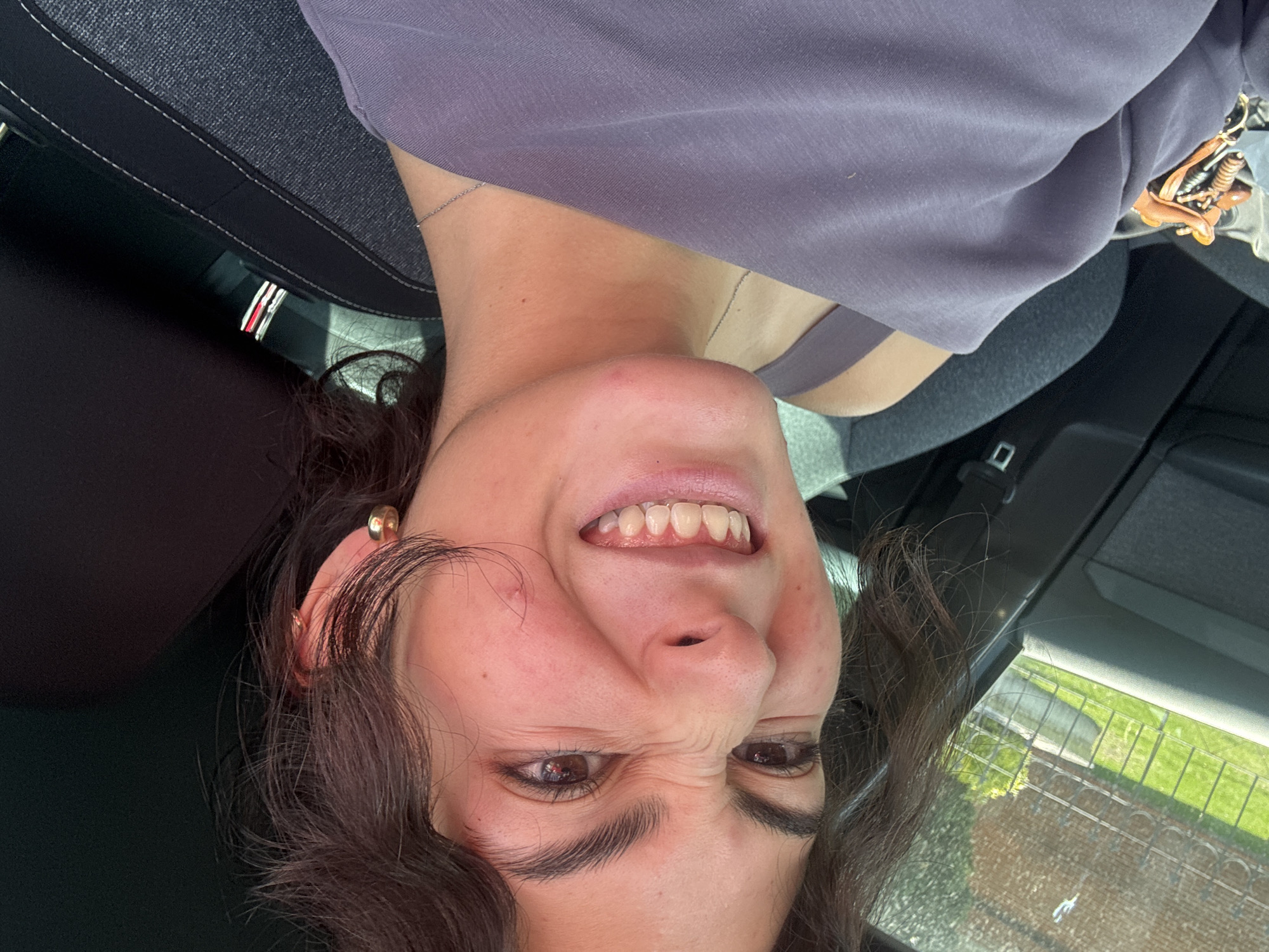 A young woman is captured in a playful selfie inside a car, presented from an inverted perspective. She has dark, curly hair and dark brown eyes, visible below expressive, prominent eyebrows. Her face is scrunched up with a broad, toothy smile, indicating a strong humorous or joyful emotion. She is wearing a light purple long-sleeved top and a thin silver necklace. A gold-toned ear cuff or earring adorns her right ear.
The setting is the interior of a car, featuring dark grey fabric seats and black panels. Through the car's window on the right side, a blurry outdoor scene is visible, depicting a green grassy area, a fence or wall, and a distant light-colored building, all bathed in daylight. Various small items, potentially keys or trinkets, are partially visible in the upper right corner of the frame. A car seatbelt buckle is also seen in the lower right. The overall impression is a casual, spontaneous moment in San Bernardino Tlaxcalancingo, Mexico. No visible text is legible.