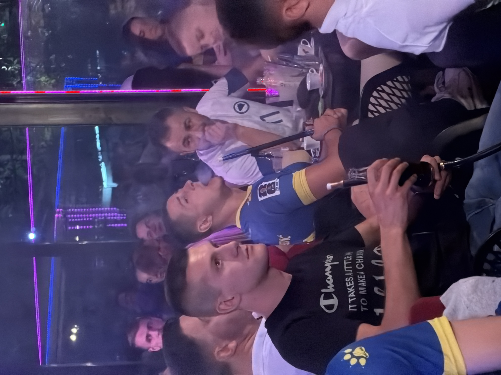 The image depicts an indoor scene, possibly a cafe or lounge, with several individuals seated around tables. The environment is dimly lit, with neon lights in blue and purple colors illuminating the background. In the foreground, three men are prominently visible. The man on the left wears a black t-shirt with a "Champion" logo and text. The man in the center wears a blue and yellow jersey with a FIFA logo, and he is holding a hookah pipe, appearing to exhale smoke. The man on the right wears a white t-shirt with a crest. Several tables are present, with drinks, cups, and an ice bucket on them. The overall atmosphere suggests a social gathering. The image is identified as being taken in Travnik, Bosnia and Herzegovina.