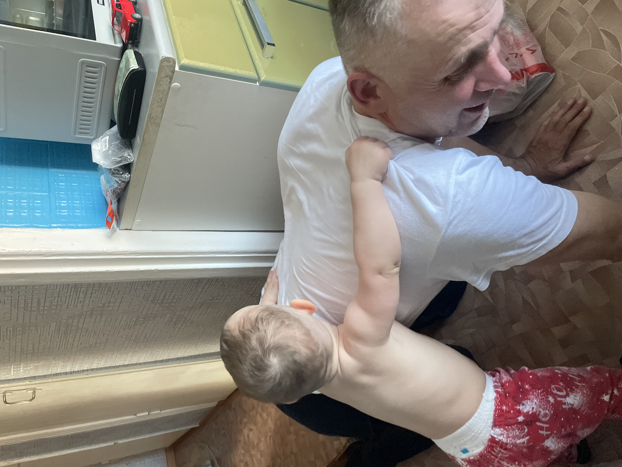 A man wearing a white t-shirt is lying on a brown surface, while a baby in a diaper and red and white pants is reaching for him. The location appears to be indoors, near a counter with cabinets and appliances. No specific details indicate the city of Siva, Russia.