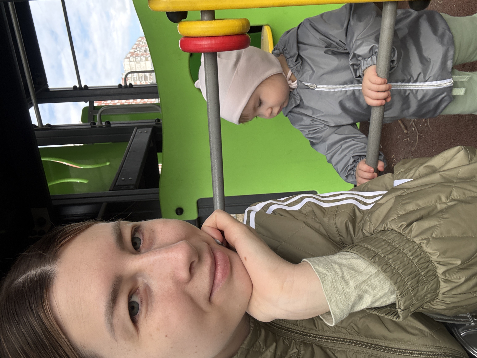 The image shows a woman with fair skin and light brown hair in a green Adidas jacket with white stripes. She is lying down and looking at the camera, with her hand resting on her chin.
Next to her is a toddler wearing a light pink hat and a gray jacket with a white zipper. The toddler is standing on a playground structure, holding onto a gray pole.
The playground structure is green, with yellow and red elements. The setting is outdoors, with a blurred building visible in the background. Based on the architecture, it is possible that the location is in the city of Moscow, Russia.