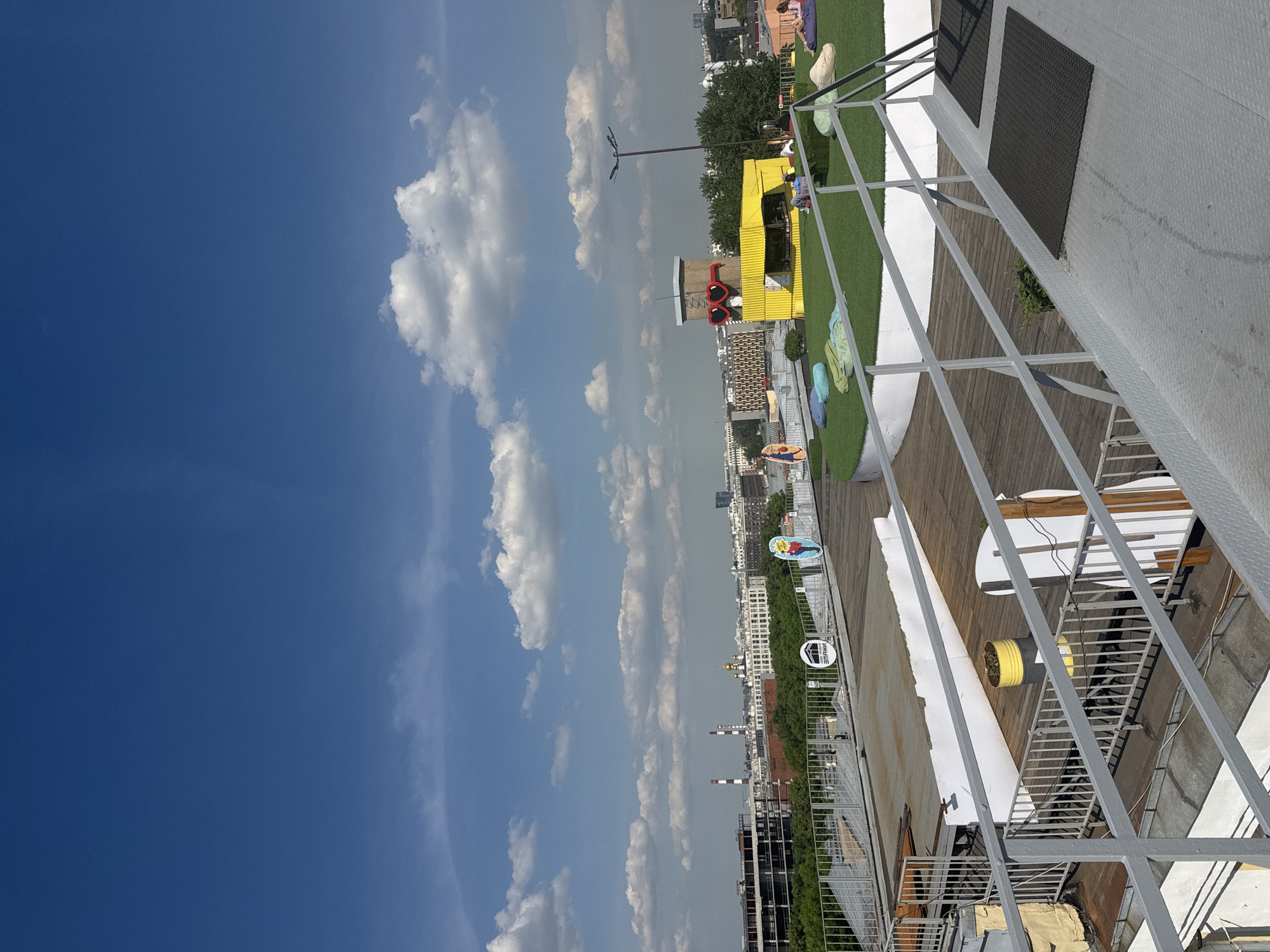 The image captures a view of a rooftop terrace in Saint Petersburg, Russia, on a bright, partly cloudy day. In the foreground, a metal walkway with a railing leads upwards, bordered by a section of wooden decking and a low white wall. A yellow and grey barrel with a plant sits on the wooden deck. Further into the scene, a circular area with artificial turf is visible, furnished with several beanbag chairs. A yellow, booth-like structure with large red heart-shaped glasses perched on top stands on the turf, resembling a quirky kiosk or bar. A lamppost stands nearby. In the background, a cityscape unfolds with various buildings, including structures with domes, industrial chimneys, and apartment blocks, all set against a vibrant blue sky dotted with white clouds. A few people are scattered on the artificial turf, relaxing on the beanbag chairs.