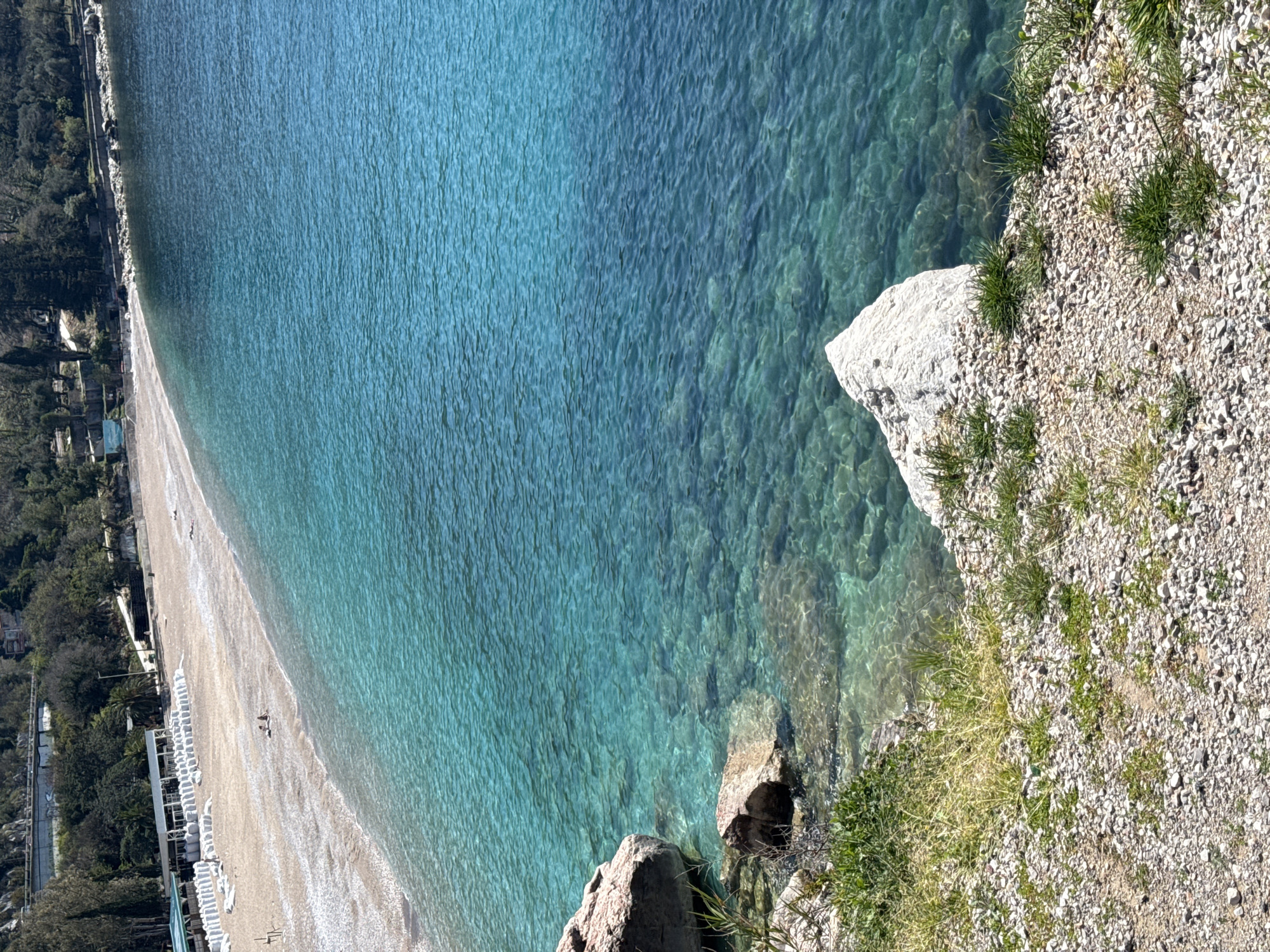 The image captures a scenic view of a beach in an unknown city. The scene features the turquoise sea, a sandy beach lined with rows of white beach chairs, and a rocky shoreline covered in small stones and patches of grass. The perspective is from above, offering a clear view of the water's transparency and the texture of the shoreline. Two people are visible walking along the beach.