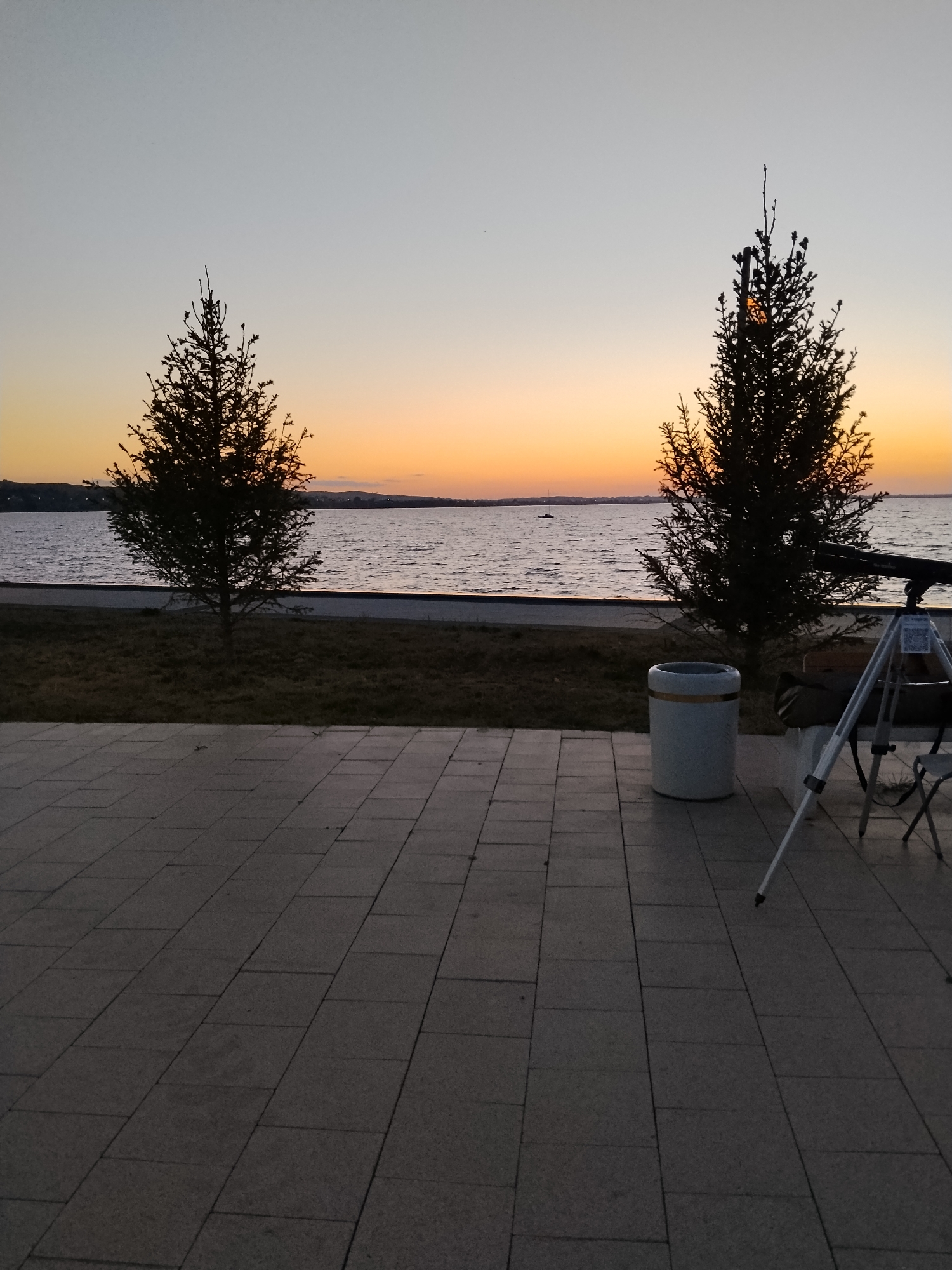 The image captures a twilight scene by a body of water, likely a lake or a reservoir, in the city of Kokshetau, Kazakhstan. In the foreground, a paved walkway made of light-colored rectangular tiles stretches across the bottom half of the frame. Beyond the walkway is a grassy area with scattered fallen leaves. Two silhouetted evergreen trees stand on the grass, one to the left and another to the right. The trees are dark against the softly colored sky, which transitions from a pale blue at the top to shades of orange and yellow near the horizon, indicating sunset or sunrise.
The body of water is calm, with gentle ripples visible on its surface. A distant, dark shoreline with hills or low buildings is discernible across the water. To the right of the frame, near the second tree, a white cylindrical trash bin with a golden band is placed on the paved area. Adjacent to the trash bin and slightly further back, a telescope is set up on a tripod. A dark chair is partially visible behind the telescope. The overall atmosphere is serene and quiet, suggesting an evening or early morning setting. The lighting is dim, consistent with twilight. There are no people visible in the image.