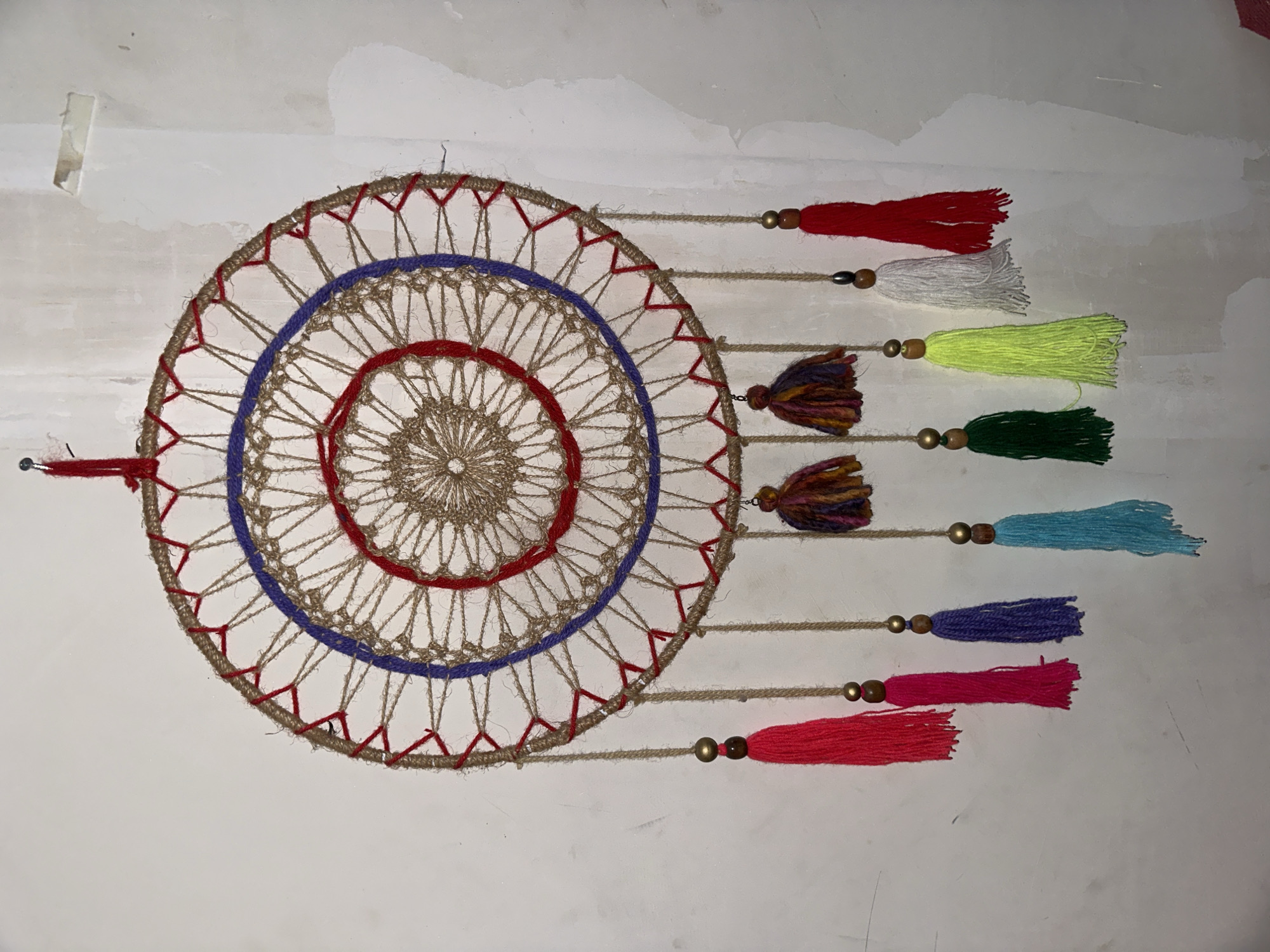 A handcrafted dreamcatcher hangs against a white wall with visible imperfections. The dreamcatcher features a circular frame with an outer layer of brown twine accented with red yarn at intervals. Inside, there are concentric circles of blue and red yarn, with a tightly woven beige center resembling a doily. Seven strands hang from the right side of the dreamcatcher. Each strand is composed of brown twine, adorned with a wooden bead and terminating in a brightly colored tassel. The tassels are red, white, yellow, dark green, light blue, purple, and pink. On the left side of the dreamcatcher, a single red strand extends outward. This scene is from the city of Arica, Chile.