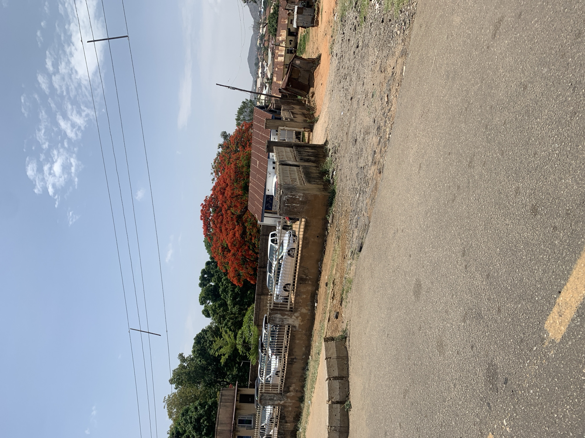 The image depicts an outdoor scene in Jos, Nigeria. A paved road occupies the foreground and right side. The road has a pale yellow paint stripe on its right edge. A concrete curb borders the road, beyond which lies a rough, dirt and gravel area.
A low, aged concrete wall runs along the edge of this area, parallel to the road. Behind the wall is a row of parked white cars and what looks like a possible retail automotive business. A vibrant red-flowered tree is present, likely a flamboyant tree. Other trees with green foliage are visible.
Behind the cars and trees are buildings, some appearing older with corrugated metal roofs. A hillside can be seen in the background. Power lines run across the upper left of the image. The sky is light blue with scattered clouds.