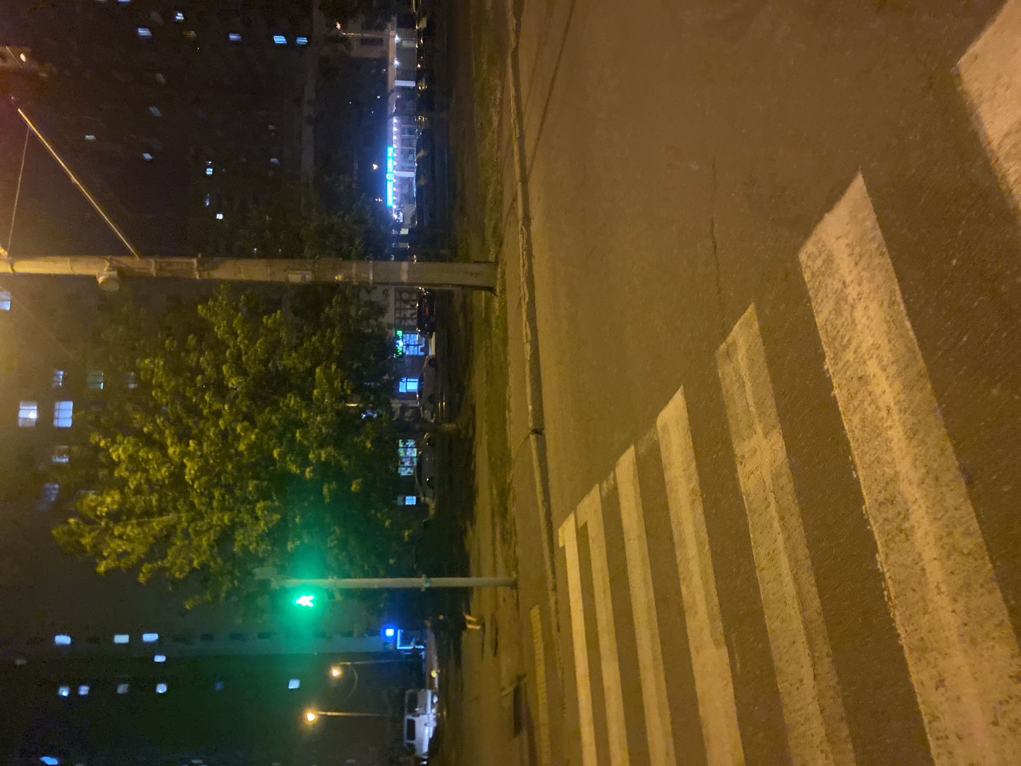 The image depicts a nighttime scene in a city, likely Kyiv, Ukraine, given the context. A pedestrian crossing, marked by white stripes on asphalt, dominates the foreground. Beyond the crossing, a street light illuminates the area, casting a yellowish glow. A traffic light, displaying a green pedestrian symbol, is visible on the right side of the image, indicating it is safe for pedestrians to cross. A lone figure, appearing to be a person in dark clothing, stands on the sidewalk near the traffic light. In the background, several multi-story apartment buildings with lit windows are visible, along with trees and other urban structures. The overall ambiance is that of a typical urban environment at night, with artificial lighting illuminating the scene.