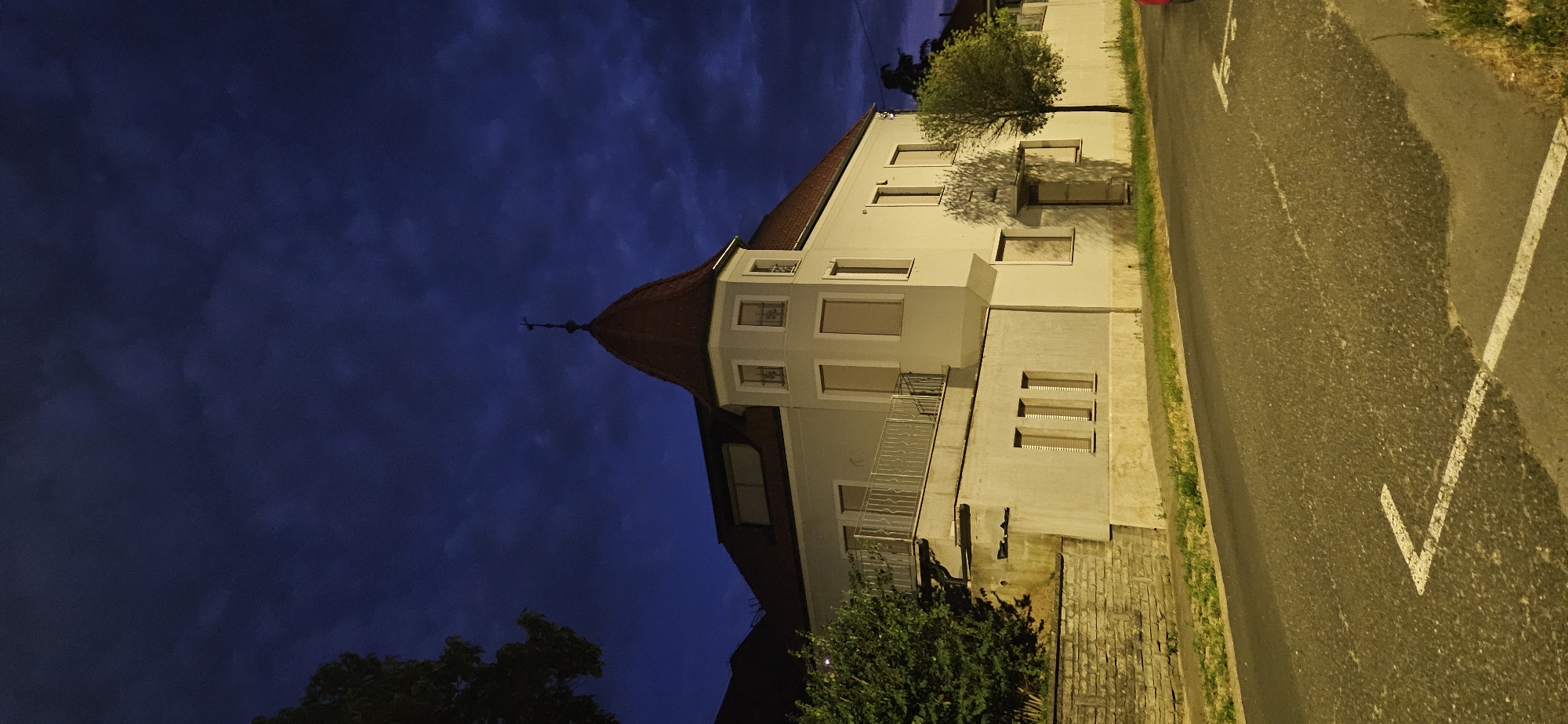 The image shows a multi-story residential building with a distinctive turret, seen at dusk or night. The building is light-colored with a red tiled roof. The sky is a deep blue with dark, textured clouds. A paved road runs in front of the building, with white road markings visible, including a chevron pattern. There is grass and a stone wall along the side of the road. A small tree with green leaves stands to the right of the building. The overall impression is that of a quiet, possibly residential street in a European town.
Specifically, this image depicts the building located at **32 Széchenyi utca, Hévíz, Hungary**. This particular building is known for its unique architectural style, featuring the prominent turret that gives it a castle-like appearance. The presence of the road, pavement, and typical vegetation suggests an urban or suburban environment within the city. The time of day, indicated by the dark sky and artificial lighting on the building (implied by the illumination of its facade), suggests an evening scene. No people or specific actions are visible in the image, focusing solely on the architecture and its surroundings.