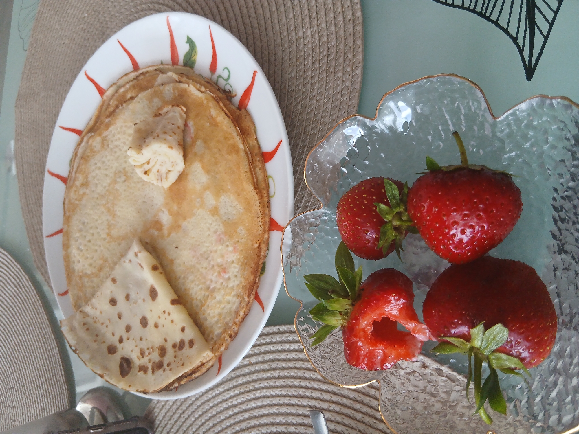 A stack of thin, golden-brown crepes, with one crepe folded into a triangle on top, rests on a white plate with a decorative red and green border. A small piece of something white and fluffy, possibly cream cheese or sour cream, is placed on the crepes. Below the plate, a clear, textured glass bowl holds several ripe strawberries, with one strawberry partially eaten and showing a hollow center. The scene is set on a table with a woven placemat. The image appears to be an indoor still life, likely a home setting in the city of Chaikovsky, Russia, given the context of the request.