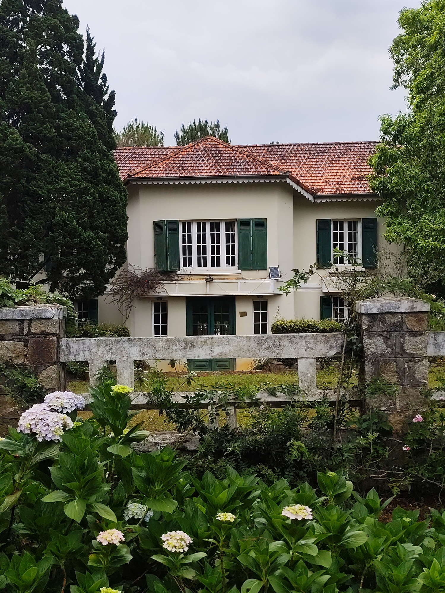 The image depicts a two-story building in Da Lat, Vietnam, characterized by its cream-colored exterior, a red tile roof with decorative edging, and dark green shutters on the windows. The building has a symmetrical facade, with a central entrance and multiple windows. A solar panel is visible mounted below a window. The building is surrounded by lush greenery, including trees and flowering plants, particularly hydrangeas in the foreground, which partially obscures a low stone and concrete fence. The sky is overcast, suggesting a cloudy or rainy day. The overall scene conveys a sense of tranquility and the aesthetic of a traditional Vietnamese home.
