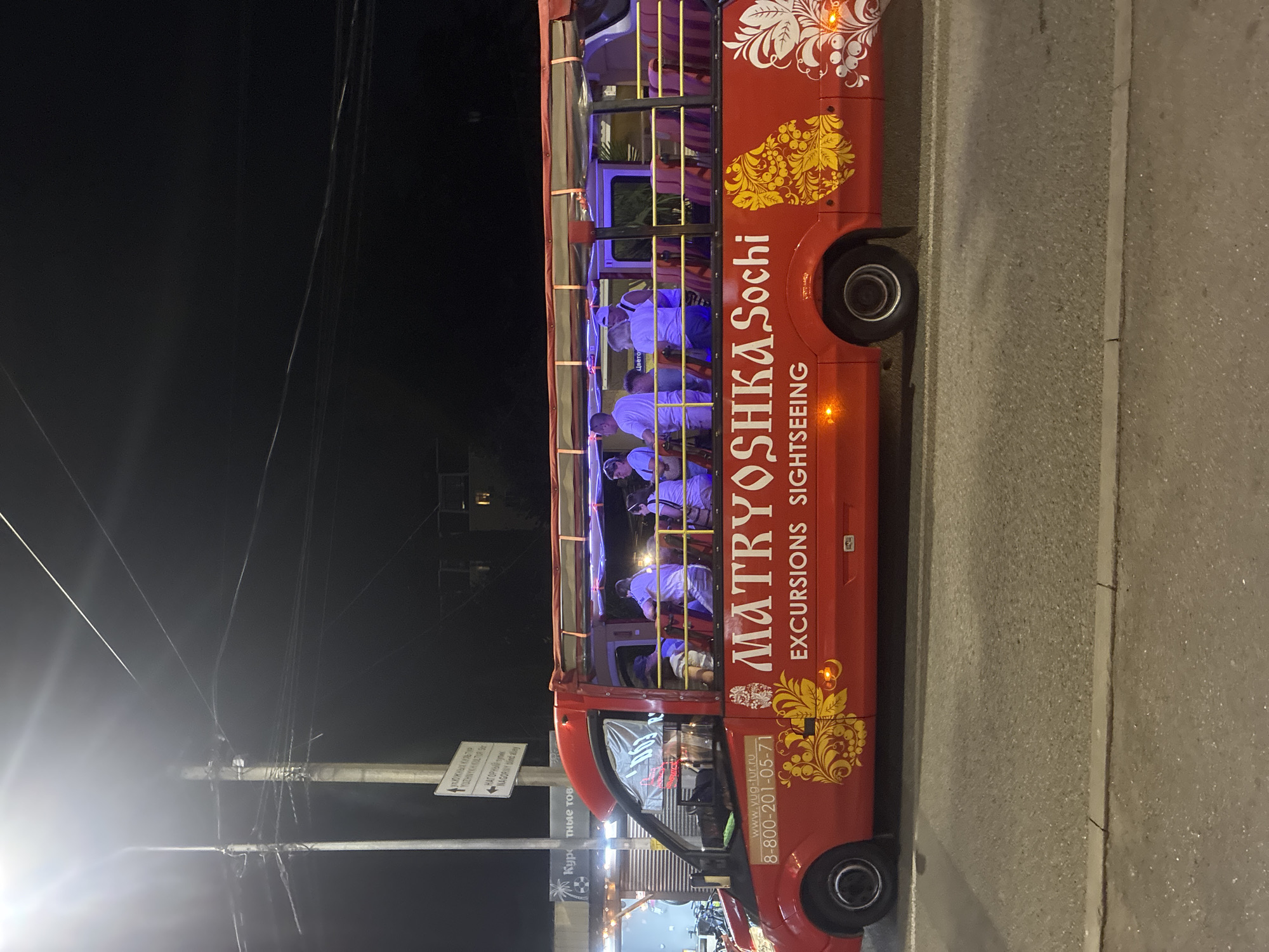 The image shows a red open-top tour bus, branded "MATRYOSHKA Sochi EXCURSIONS SIGHTSEEING," driving on a street at night. The bus has several passengers visible, some standing and others seated. The bus is decorated with ornate yellow and white patterns. On the side of the bus, a phone number and website are visible: "8-800-201-05-71" and "www.yug-tur.ru". In the background, streetlights and buildings are dimly visible. The street appears to be paved, and there is a curb separating the road from a sidewalk. The scene is illuminated by artificial light, suggesting it is nighttime. This scene is consistent with the city of Sirius, Russia, known for its tourist attractions and evening activities.