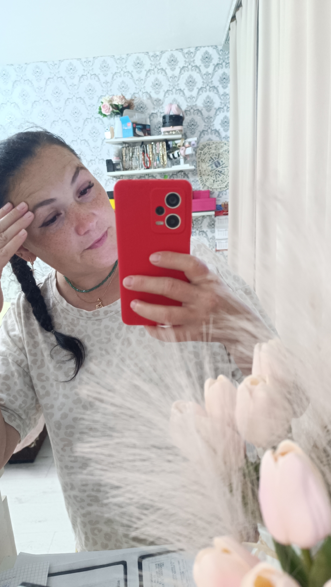 A woman with dark hair in a braid is taking a mirror selfie in what appears to be a dressing area. She is wearing a light-colored t-shirt with a leopard print. She is holding a red smartphone in her right hand, taking the photo. Her left hand is on her forehead. Behind her, a shelf is filled with various items, including jewelry and decorative objects, set against a patterned wallpaper. In the foreground, a blurry arrangement of light-colored tulips and fluffy ornamental grass is visible, suggesting the presence of a mirror and a decorated vanity or table. The background curtain on the right is light-colored and opaque. The woman is wearing a green beaded necklace and a small pendant necklace.