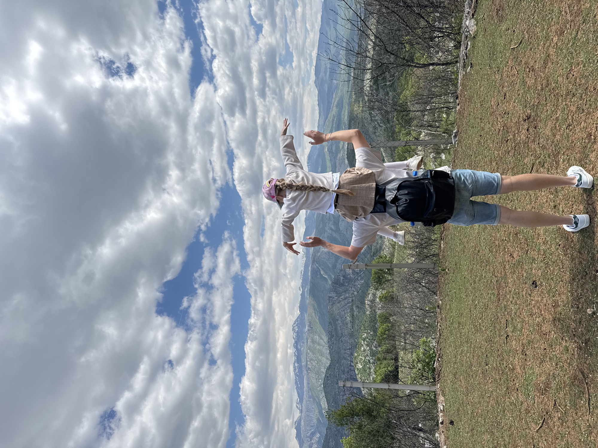 A medium shot shows a person facing the sky, with their back toward the camera. The setting appears to be a mountaintop overlooking a valley in Gornje Mrke, Montenegro. The person is wearing blue jean shorts, a light colored top, and white tennis shoes. They have a backpack on and a pink hat with a blonde braid coming out of it. The person's arms are raised. The sky is filled with white, puffy clouds with patches of blue sky in between. In the background, a mountain range stretches across the horizon. In the foreground, there is a patch of grass with some trees and shrubs. A wire fence is visible in the middle ground.