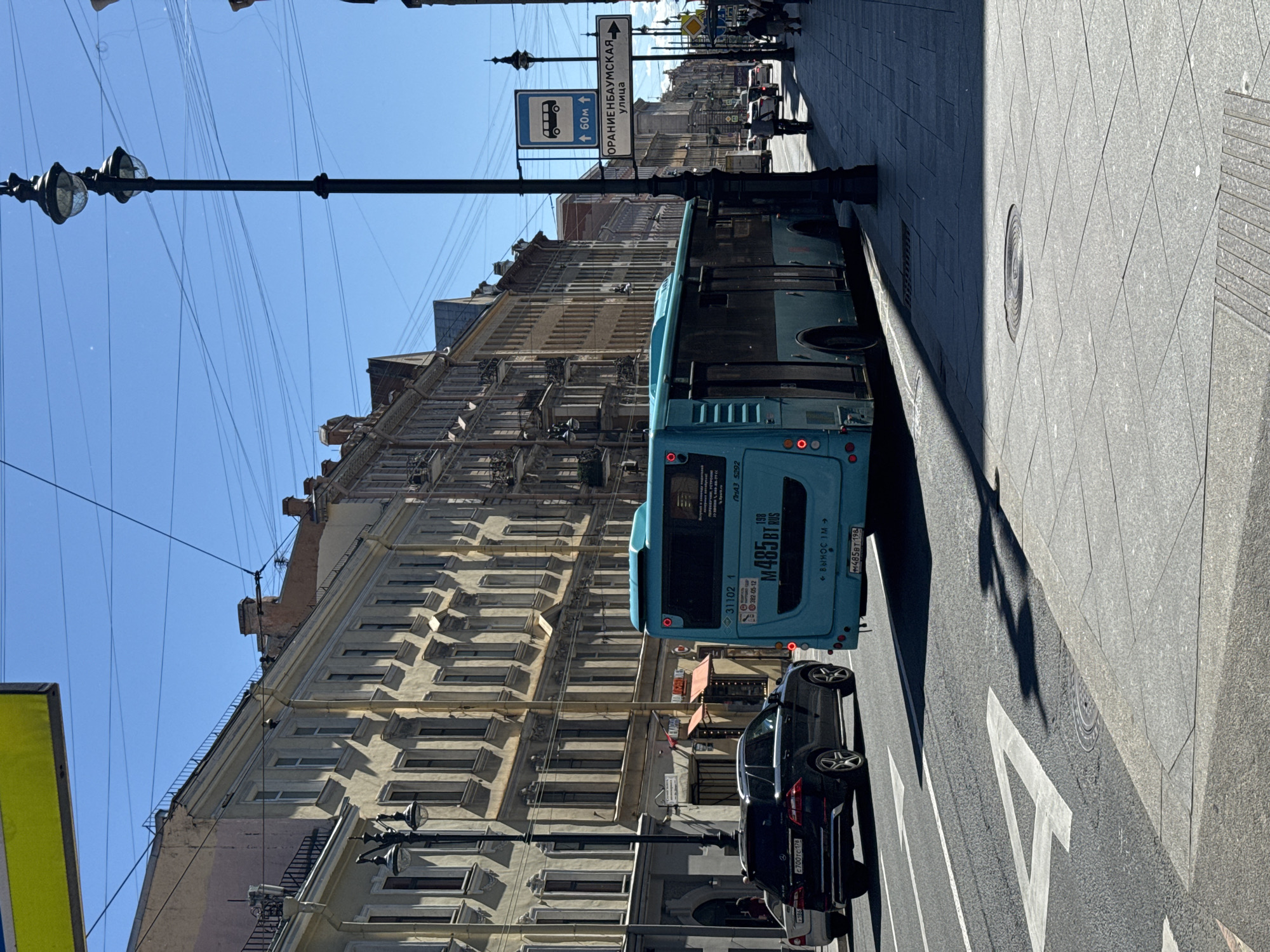This media file captures an outdoor urban scene dominated by a light blue bus. The bus is the most prominent object, seen from the rear and partially from the side, with its license plate and route information clearly visible. Next to the bus, a dark sedan is parked, also showing its license plate. The street is paved with grey tiles, and white lane markings are visible on the asphalt.
A tall, ornate street lamp stands on the left side of the image, with its classic design contrasting with the modern vehicles. Overhead, a complex network of electrical wires crisscrosses the bright blue sky, suggesting a busy urban environment.
In the background, a long, multi-story building with numerous windows lines the street. The architecture of the building appears historic, with decorative elements suggesting an older European city. A street sign with Cyrillic text and directional arrows, along with a bus stop sign, are visible above and to the left of the bus, providing location cues.
The lighting indicates it is daytime, with strong sunlight casting shadows. The clear blue sky suggests fair weather. No people are clearly discernible as distinct subjects, though some figures might be present in the distance. The overall impression is a snapshot of daily urban life, likely in a city with a significant public transportation system and a mix of old and new infrastructure. The visible text includes Russian street names and vehicle identification numbers.
