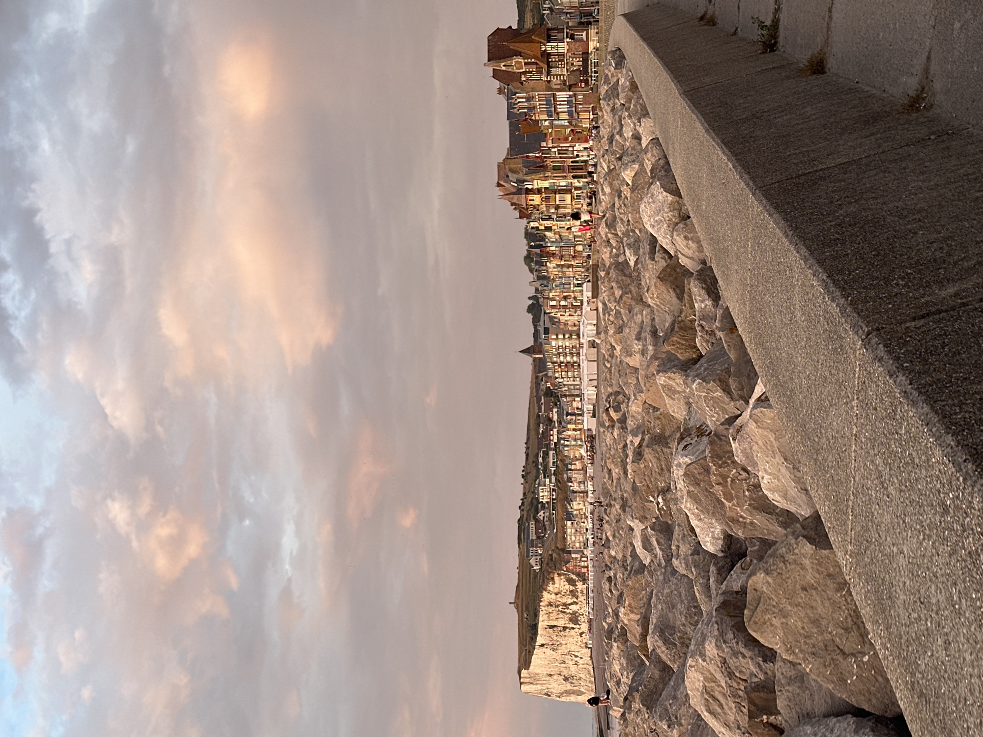The image depicts a coastal scene in Le Tréport, France, characterized by a rocky breakwater in the foreground and a clifftop town in the background. The breakwater, composed of large, irregularly shaped rocks, extends into the frame from the left, forming a barrier along the coastline. A concrete walkway borders the breakwater on the right.
In the middle ground, a sandy beach is visible, lined with beach huts or shelters. Several people are present on the beach and near the water's edge, appearing to be enjoying the seaside. The town of Le Tréport is situated atop a prominent white cliff that dominates the left side of the background. This cliff face is steep and imposing. Buildings of the town, featuring colorful facades and distinctive architecture, stretch along the clifftop and down towards the coast.
The sky above is filled with clouds, tinged with soft pink and orange hues, suggesting either sunrise or sunset. The overall lighting is warm and atmospheric. A lone figure stands on one of the large rocks in the foreground, looking out towards the sea and the town. The environment is clearly a coastal resort town, with elements of natural landscape and urban development coexisting.