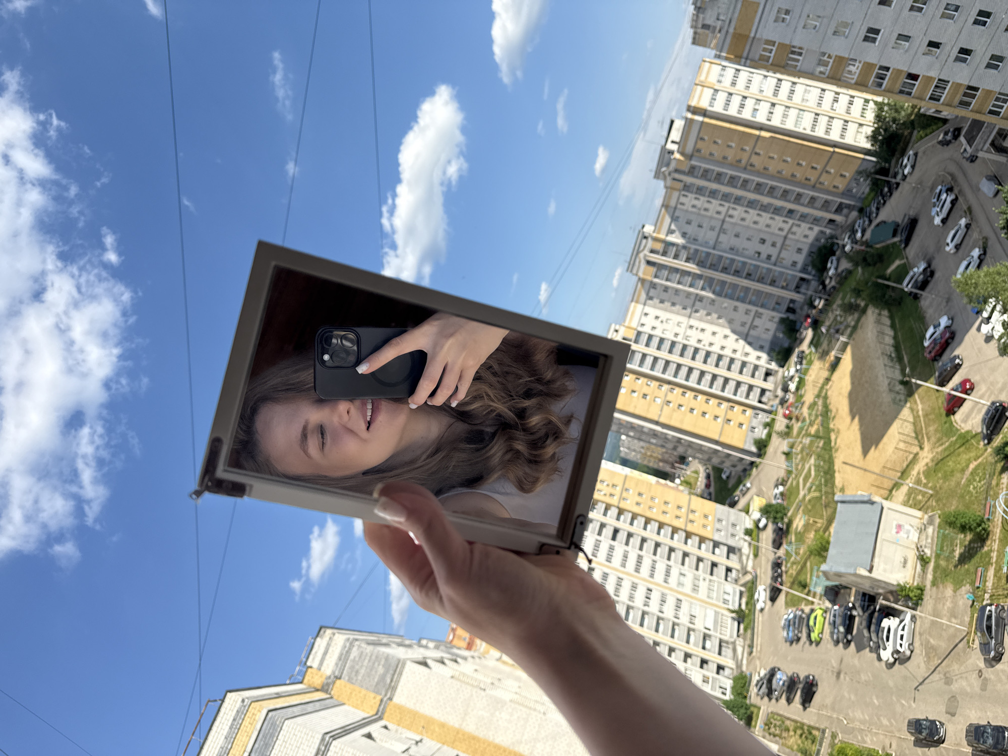 A young woman is taking a mirror selfie outdoors, holding a rectangular mirror in her right hand, which reflects her face and upper body. She is looking at the phone she is holding in her left hand, with which she is taking the photo. The background consists of a bright blue sky with scattered white clouds, and in the middle ground, a cityscape with residential buildings. The buildings appear to be multi-story apartment blocks, typical of urban areas. The woman has light brown hair and is wearing a white top. The mirror is framed in a dark material, and the reflection shows the back of her phone, which has multiple camera lenses. The overall lighting suggests it is daytime, likely sunny. The city appears to be Vladimir, Russia, given the architectural style of the buildings and the presence of overhead wires, which are common in Russian cities.