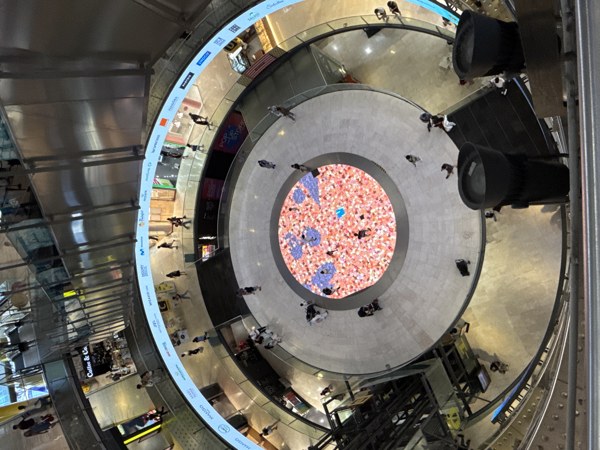 The image shows a high-angle, wide shot of the interior of a multi-level shopping mall in Barcelona, Spain. The mall features a circular atrium with a large, illuminated floor display in the center, filled with a mosaic of pink and blue lights, resembling an abstract floral pattern. Several people are visible walking on the different levels of the mall, with some gathered around the central display. The architecture is modern, with curved balconies, metal railings, and large glass panels. Stores with illuminated signage are visible along the upper levels, including "Coffee & Co.", "MANGO", "MINT & TERRA", "MISAKO", "NESPRESSO", and "PANDORA". Escalators are present, and the overall environment appears to be well-lit and bustling with activity. The perspective suggests the photo was taken from an upper floor or a higher vantage point looking down into the atrium.