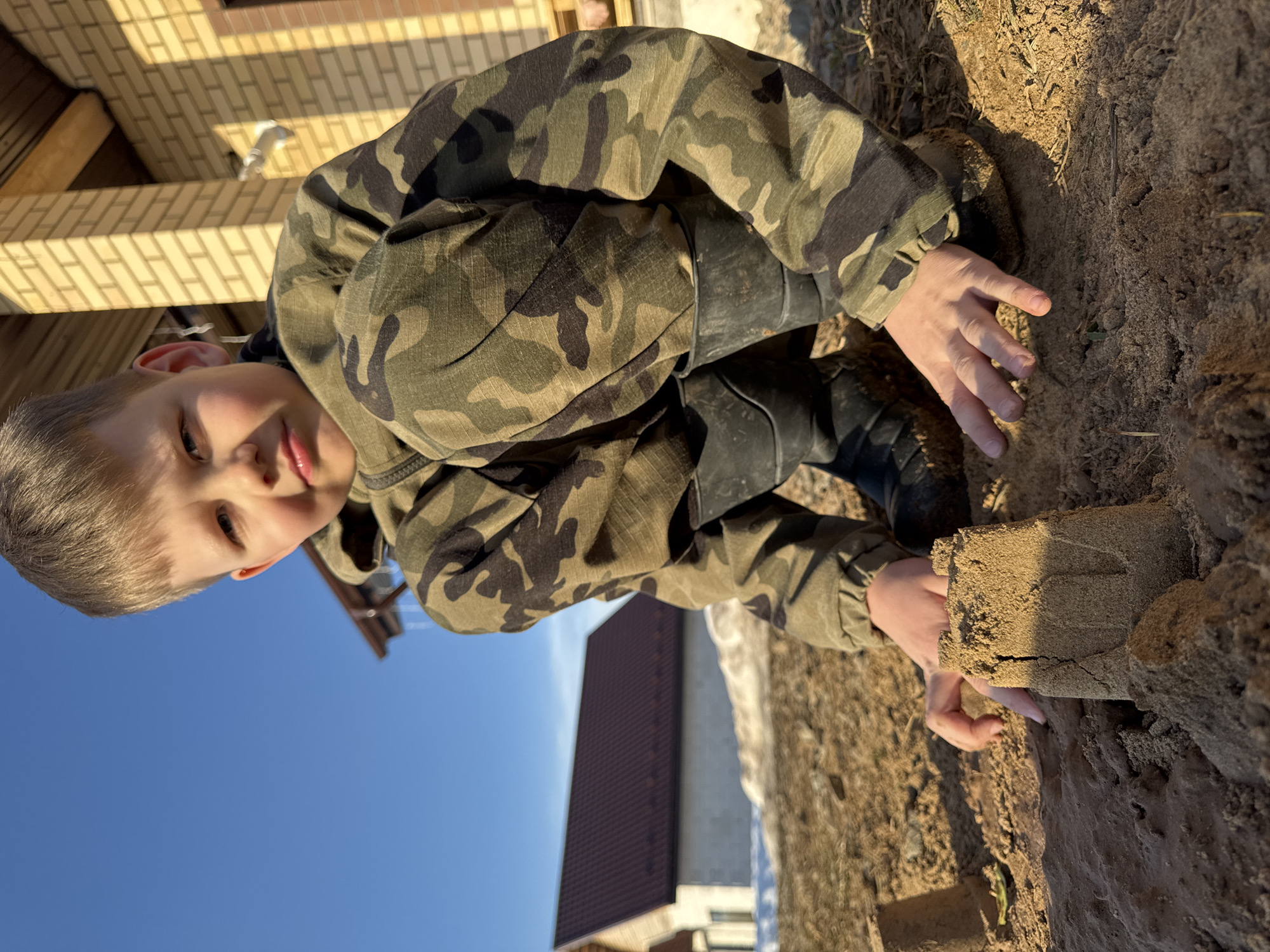 An outdoor daytime scene in Gornyi (Dvurechenskoe S/P), Russia, depicts a young boy squatting on light brown soil or sand. The boy, estimated to be between 5-10 years old with short, light brown hair, is wearing a green, brown, and black camouflage hooded jacket and matching pants, along with black rubber boots. He looks directly at the camera with a neutral expression. His hands are positioned in the soil near several small, rudimentary sand or dirt structures, including one roughly cylindrical shape and another resembling a small, partially cracked sandcastle in the immediate foreground. In the background to the right, a section of a light yellow brick building with a dark brown roofline and visible wooden beam is present. An outdoor light fixture is mounted on the brick wall. To the boy's left, a patch of melting snow or slush is visible on the ground. Further left and behind, a distinct white building with a prominent dark brown pitched roof stands against a clear, bright blue sky. Strong sunlight illuminates the scene, casting distinct shadows.