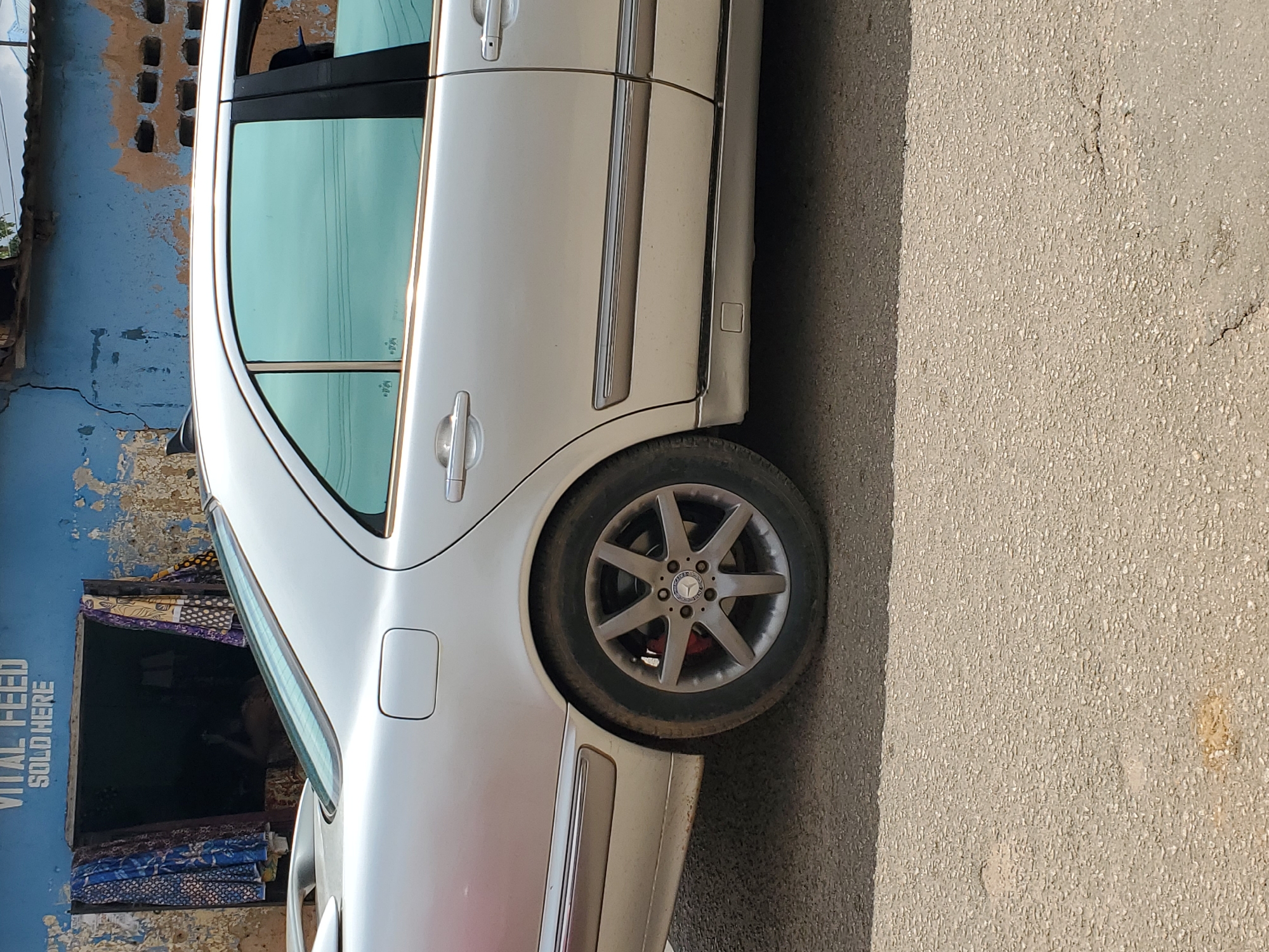 A silver Mercedes-Benz sedan is parked on a textured asphalt road in front of a dilapidated building in Jos, Nigeria. The building's blue facade is peeling and cracked, revealing weathered concrete and brickwork. Above an open doorway, white text reads "VITAL FEED SOLD HERE." Brightly patterned fabrics hang on either side of the doorway, obscuring the interior. The car's tinted rear windows reflect the sky. The rear wheel, featuring a multi-spoke alloy rim with red brake calipers visible, is prominent in the foreground. The overall scene suggests a commercial area with signs of age and disrepair.