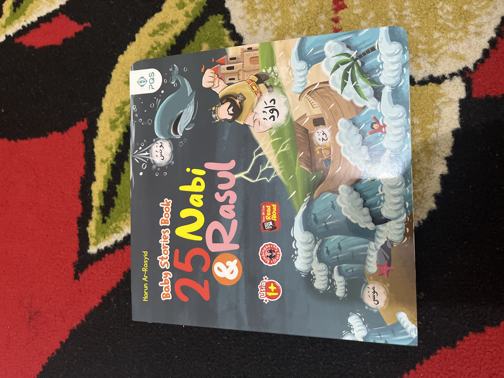 The image shows a book titled "25 Nabi & Rasul Baby Stories Book" by Harun Ar-Rasyid, placed on a red and yellow patterned carpet. The book cover features cartoon illustrations of various biblical figures and scenes, including a whale, a man labeled "Daud" (David), a boat with animals labeled "Nuh" (Noah), and a figure labeled "Musa" (Moses) amidst waves. There are also smaller labels with Arabic text and icons indicating age appropriateness and a QR code for "Read Aloud." The lighting suggests it is indoors, and the carpet pattern indicates it might be in a home environment. There is no discernible information that places the image in East Jakarta, Indonesia.