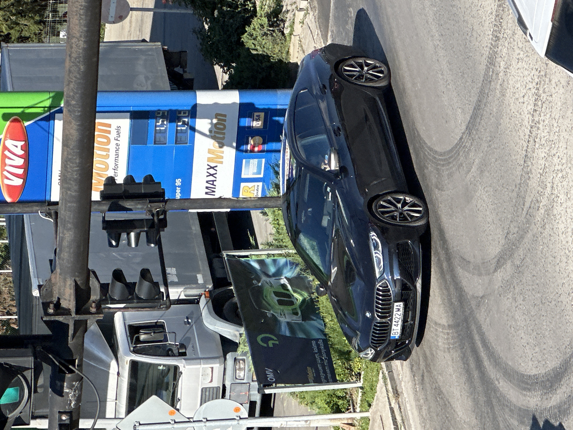A dark colored BMW sedan is driving on a paved road in Veliko Tarnovo, Bulgaria. The car has a license plate that reads "BT 4422 MA". In the background, there is a gas station with a blue and white sign that says "VIVA" and "MAXX Motion". There is also a large gray truck and a traffic signal. The road has tire marks on it, suggesting that the car may have been driven erratically. The lighting suggests it is daytime.