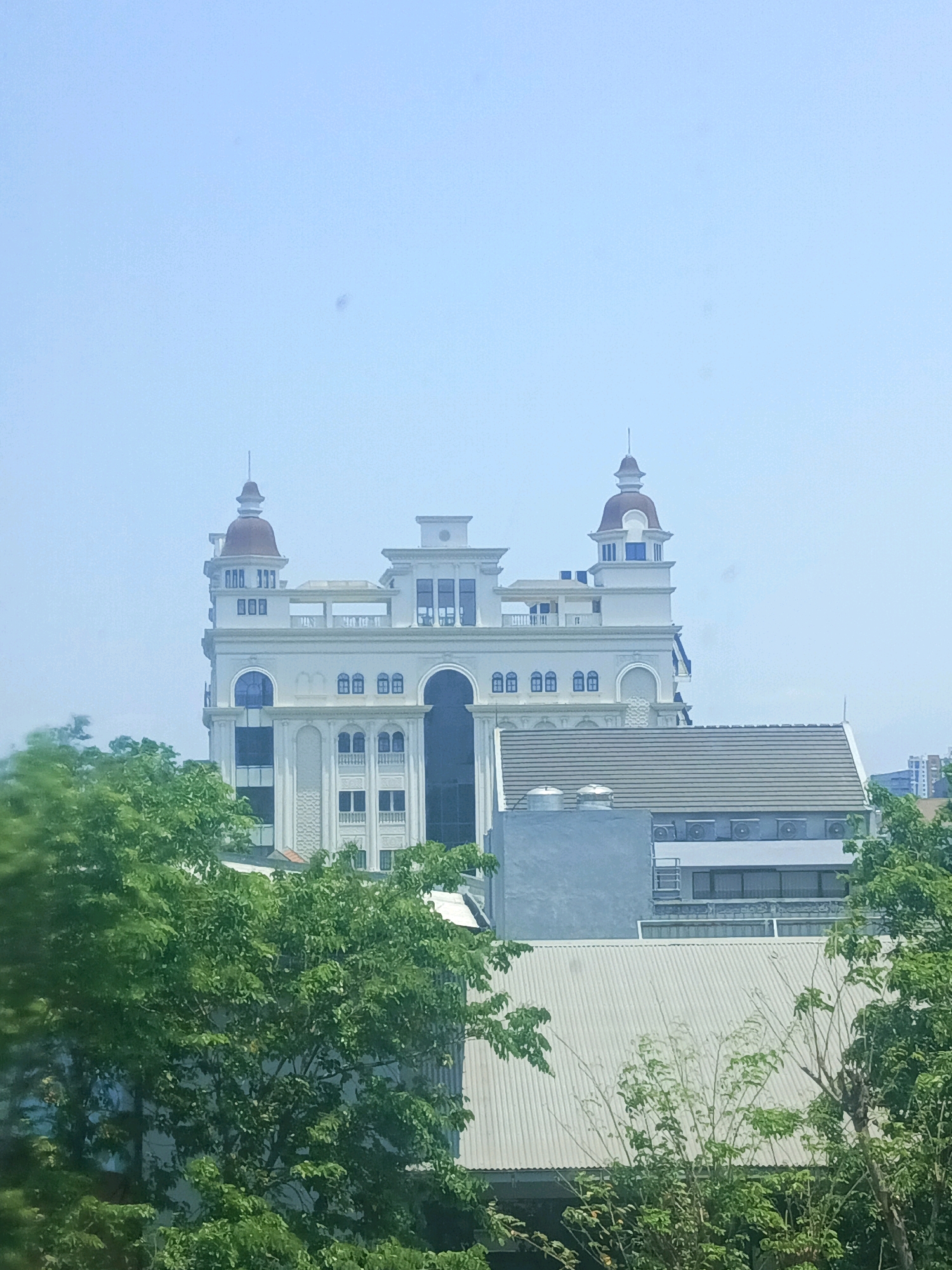 The image shows a white, ornate building with a dome-like structure on top, resembling a castle or a European-style architectural design. The building has multiple floors, with arched windows and decorative detailing. It is surrounded by lush green trees in the foreground and middle ground, with other less distinct buildings visible in the background. The sky is clear and light blue, suggesting a daytime scene. The overall impression is one of a well-maintained and perhaps historic or uniquely designed building situated in an urban environment. This building is likely located in the city of Surabaya, Indonesia.