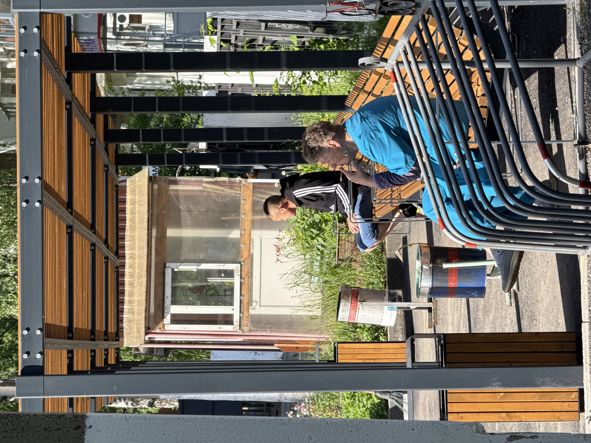 Two people, one wearing a blue shirt and another with a black and white track jacket, are seated on a bench adjacent to a metal barrier, likely engaged in conversation. A bucket sits near the bench. In the background, a structure resembling a greenhouse or shed with a transparent wall is visible. The scene seems to be located in an outdoor urban environment with greenery interspersed. The scene is located in the city of Chaikovsky, Russia.