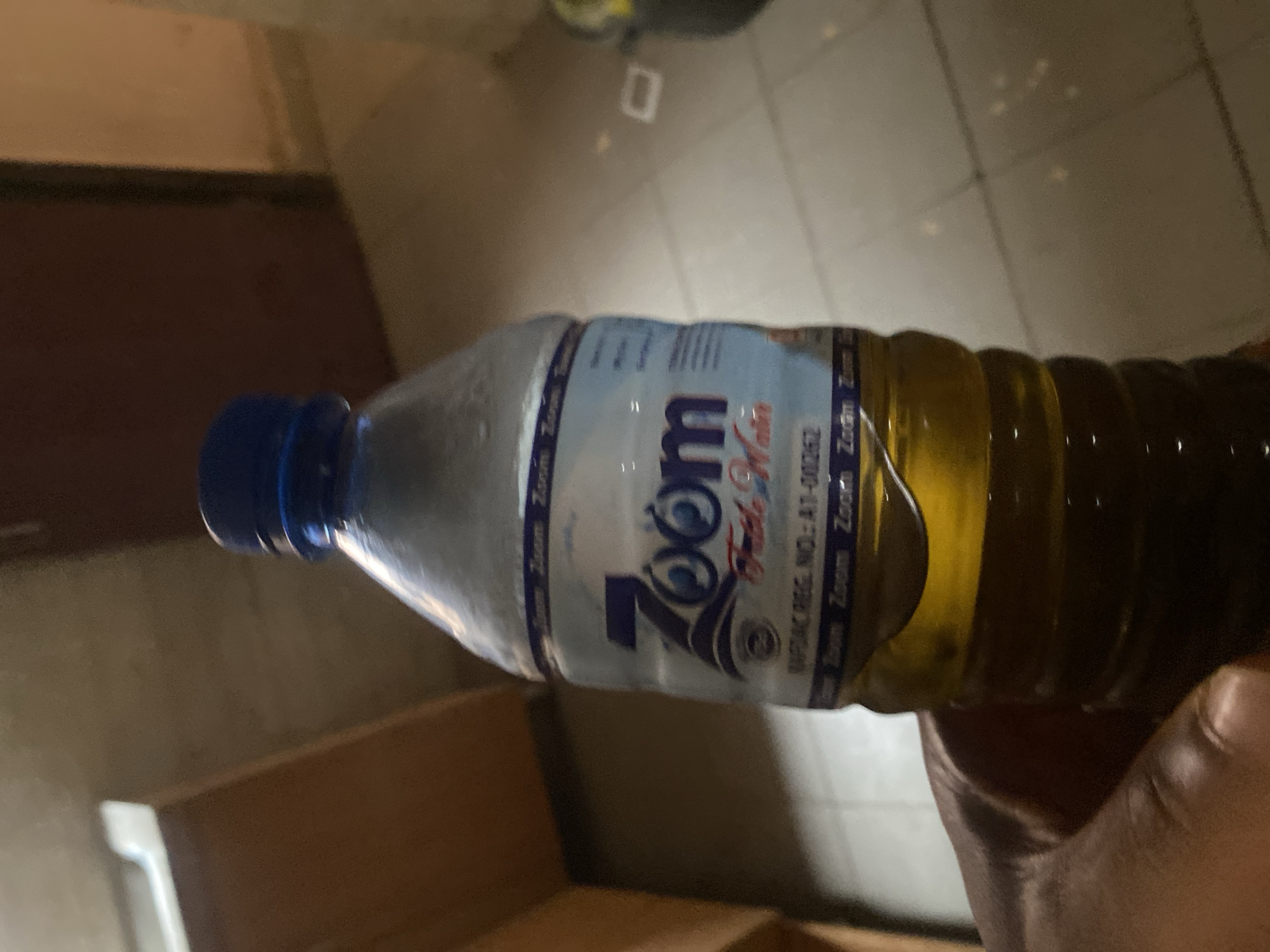 A hand holds a clear plastic bottle of Zoom brand fresh water. The bottle has a blue cap and a label with a blue and white design, featuring the word "Zoom" in large letters with two eyes. Below it, smaller text reads "Fresh Water." There is also a registration number visible on the label. The liquid inside the bottle is a yellow-brown color. The background appears to be an indoor environment with tiled flooring and wooden cabinets or furniture. The image is taken from a low angle, focusing on the bottle. The setting is described as Bulamare, Nigeria.