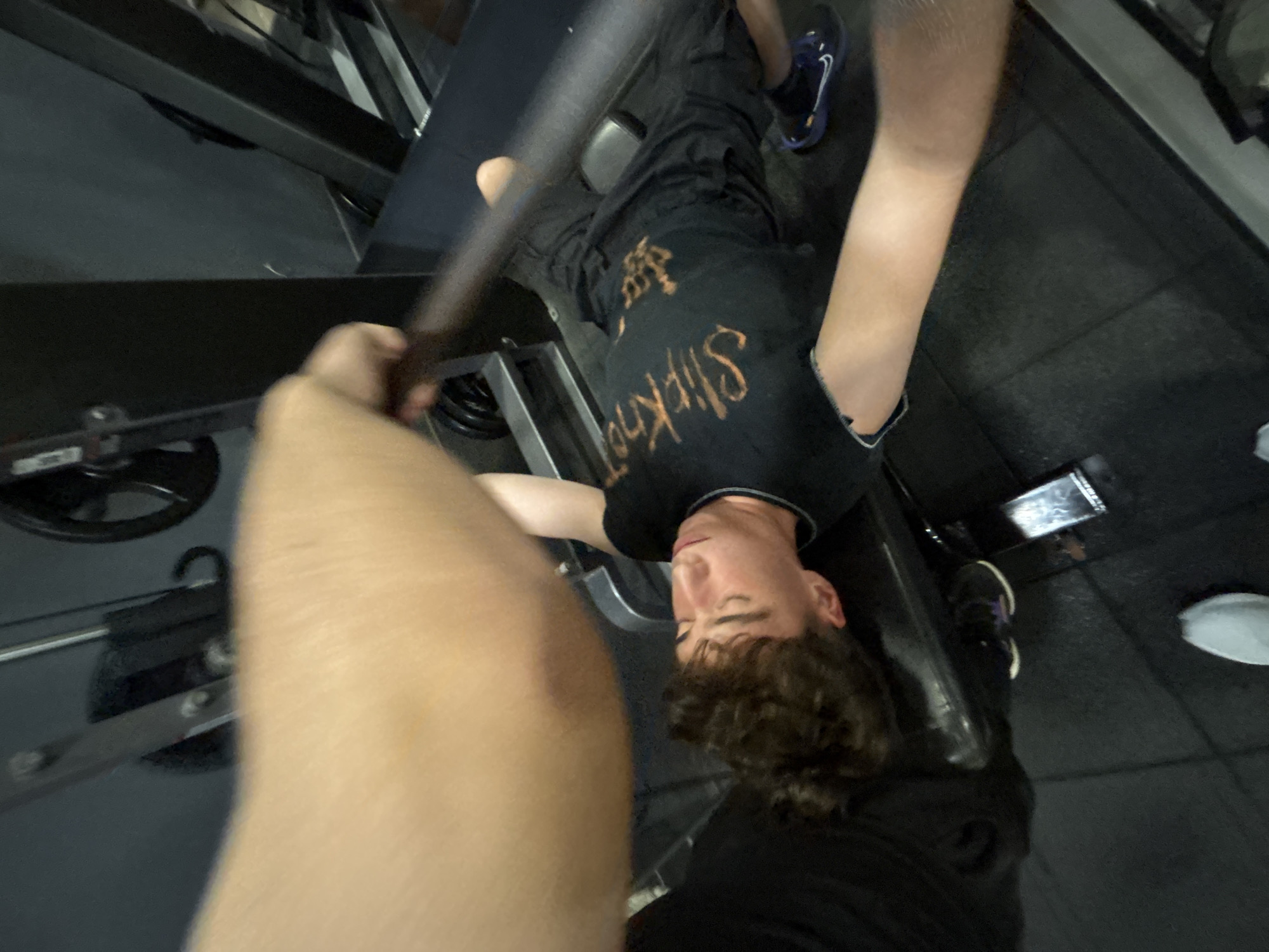 An overhead, slightly blurred shot captures a young person lying on a gym bench, seemingly in the middle of a workout. The image is oriented such that the person's head is towards the bottom of the frame, appearing upside down relative to the viewer. A light-skinned arm and hand are prominently in the foreground, possibly holding the camera or assisting with the exercise.
The subject on the bench has short, curly brown hair and a neutral expression, looking upwards. They are wearing a black short-sleeved t-shirt with the band name "Slipknot" printed in orange or brownish lettering across the chest. Dark pants and one athletic shoe with blue and white accents are visible on their left foot.
The setting is a gym, indicated by the black padded bench, a metal bar or barbell above the person's chest (partially obscured by the foreground arm), and dark rubberized flooring. A weight plate is faintly visible in the bottom left background. The overall scene suggests a workout session, likely a bench press, taking place in an indoor fitness facility in Troyan, Bulgaria.