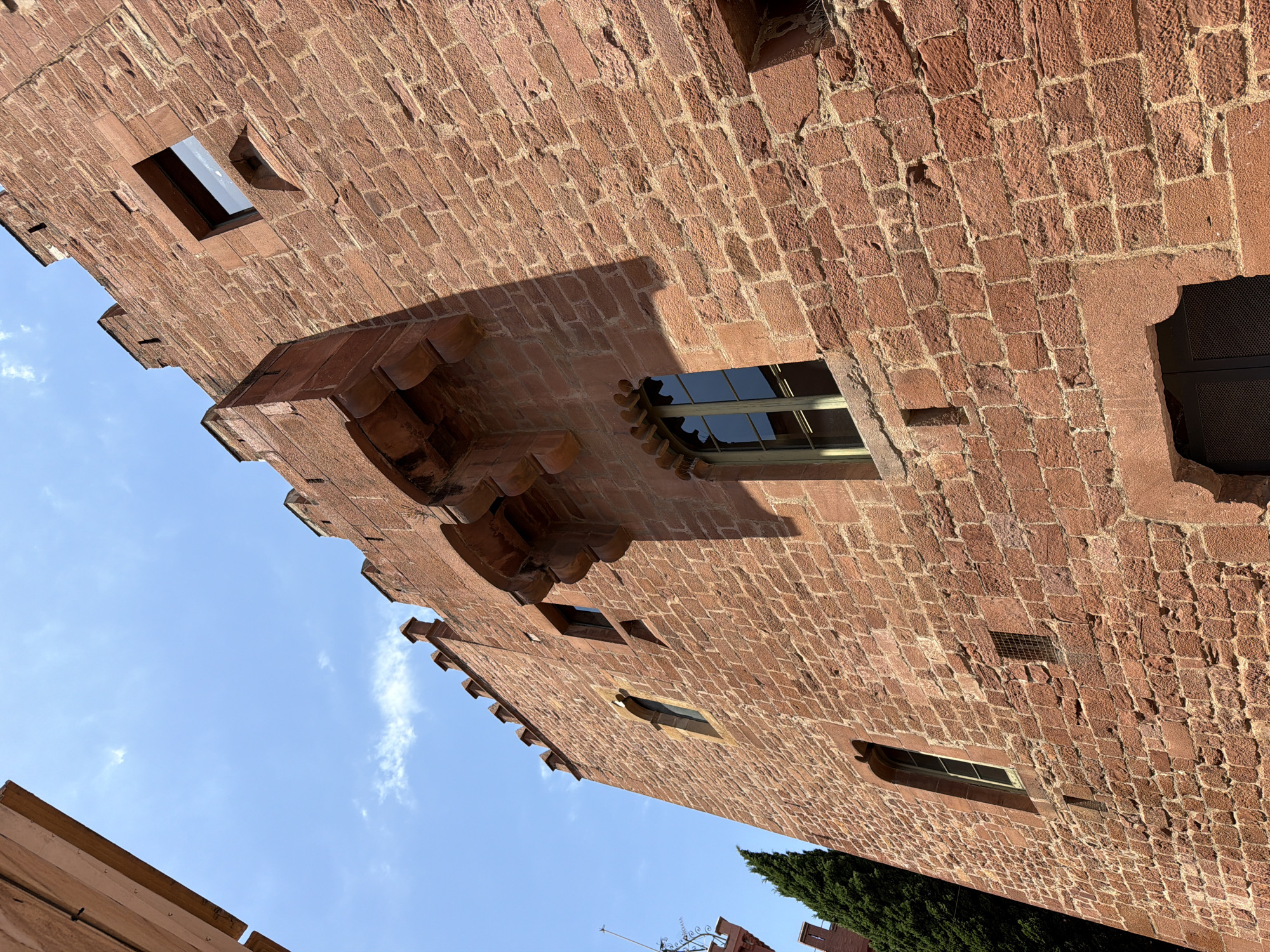 This is a low-angle, full-shot photograph of the side of a historic building, likely a castle or fortified structure, made of red sandstone blocks. The building is tall and has a crenellated roofline, suggesting defensive architecture. Several windows are visible, some with dark frames and glass reflecting the sky. A prominent feature is a projecting stone structure extending from the upper wall, supported by decorative corbels. The texture of the rough-hewn stone blocks is clearly visible, with some showing signs of age and weathering. The sky is a bright blue with a few scattered white clouds. A tall, dark green evergreen tree is partially visible on the left side of the image, and a sliver of another building made of lighter colored stone can be seen in the upper left corner. The image appears to be taken during the day, with sunlight casting strong shadows across the facade. This building is the Torre Baró de Viladecans, located in Viladecans, Spain.