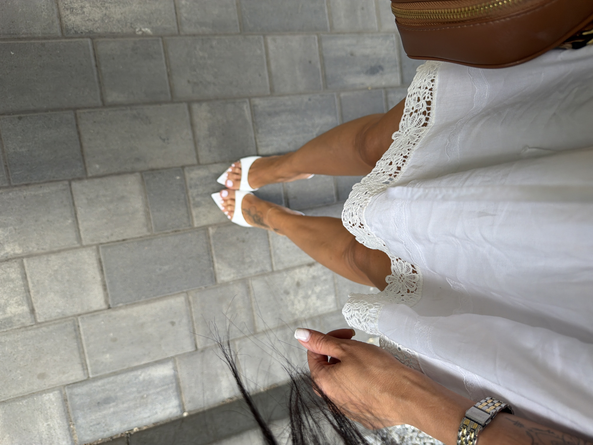 This is a high-angle, close-up shot of a person's legs and torso, standing on a grey paved walkway. The person is wearing white heeled sandals and a white dress with lace trim. A brown purse is visible on the right side of the frame. The background consists of rectangular grey pavers. There is a woman's hand with painted nails on the left side of the image, holding a section of dark hair. The scene is set in an outdoor urban environment, likely a public walkway. The specific location in Novaya Usman', Russia, cannot be determined from the visual information provided in the image.