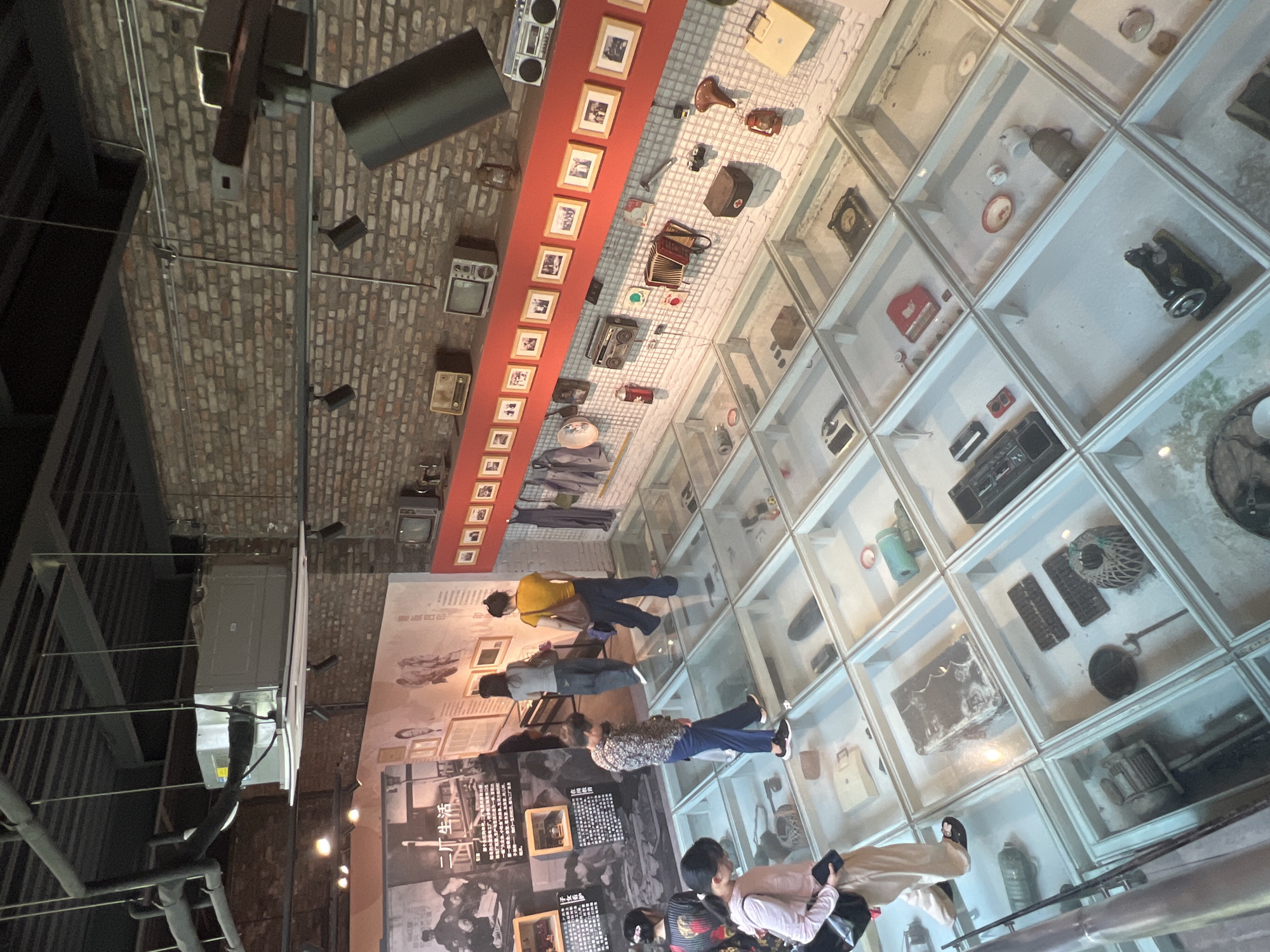 The image depicts an indoor exhibition space, likely a museum, in Yuzhong District, China. The perspective is from an elevated viewpoint, looking down at a display floor composed of glass-covered compartments. Various antique objects, including radios, a sewing machine, lanterns, and other household items, are arranged within these compartments.
Several individuals are present in the scene, interacting with the exhibit by observing the displays. One woman, wearing a pink top and beige pants, stands near the foreground, looking down at the glass floor. Another woman in a patterned shirt and dark pants is also looking at the exhibits. Further into the scene, two women in casual attire, one in a yellow shirt and the other in grey, are observing the display. The background reveals a brick wall adorned with framed photographs and several vintage electronic devices such as televisions and boomboxes displayed on a red shelf. An air conditioning unit is visible suspended from the ceiling. The overall environment suggests a historical or cultural exhibition focusing on everyday life in a past era.
