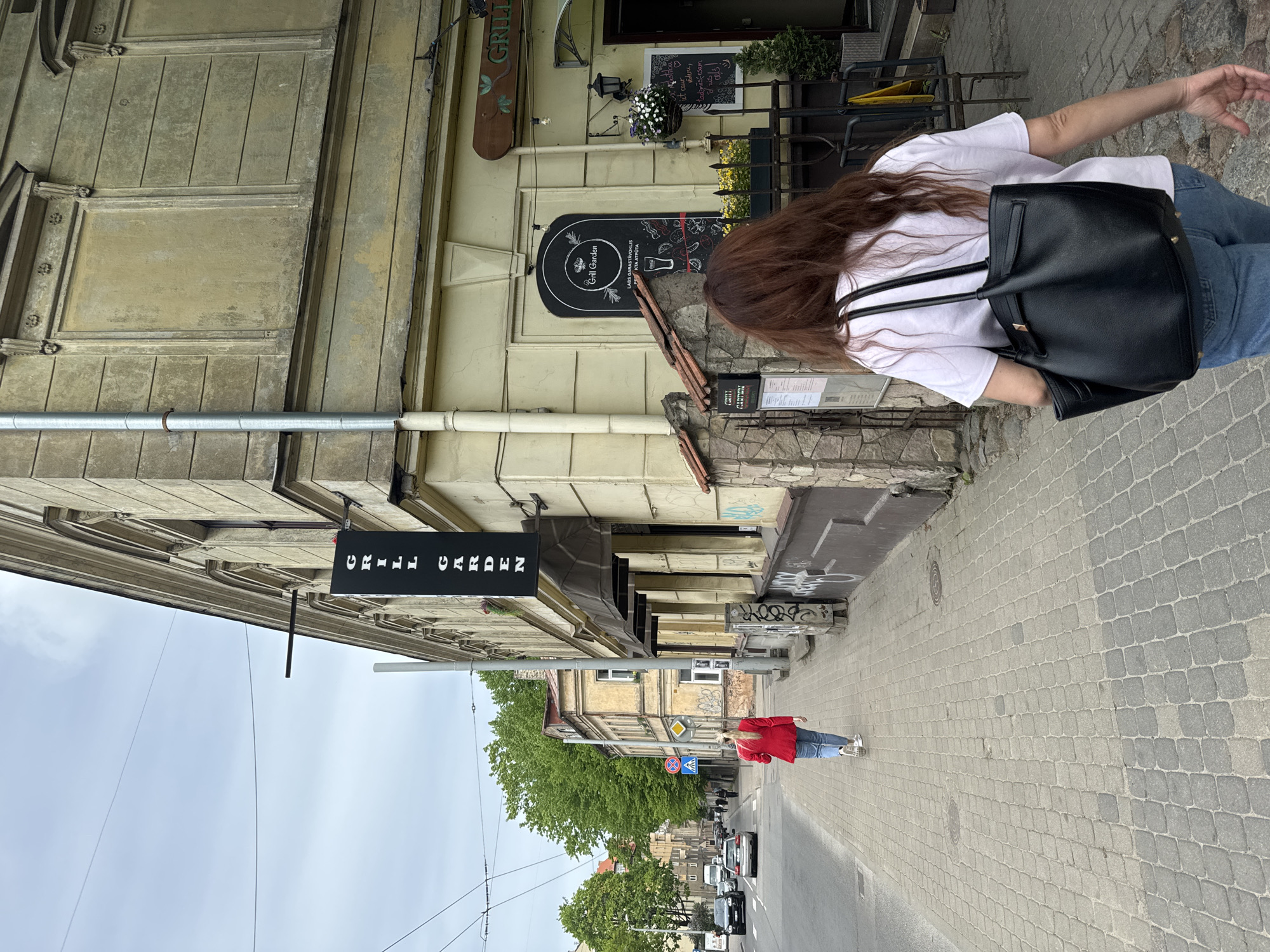 The image shows a street scene in Riga, Latvia. A woman with long brown hair, wearing a white t-shirt, blue jeans, and carrying a black leather backpack, is walking along a paved sidewalk next to a building. The building is likely a restaurant called "Grill Garden", judging by the sign hanging from the building. Another woman, wearing a red jacket and blue jeans, is walking further down the street. There are several cars and trees visible in the distance. The sky is overcast.