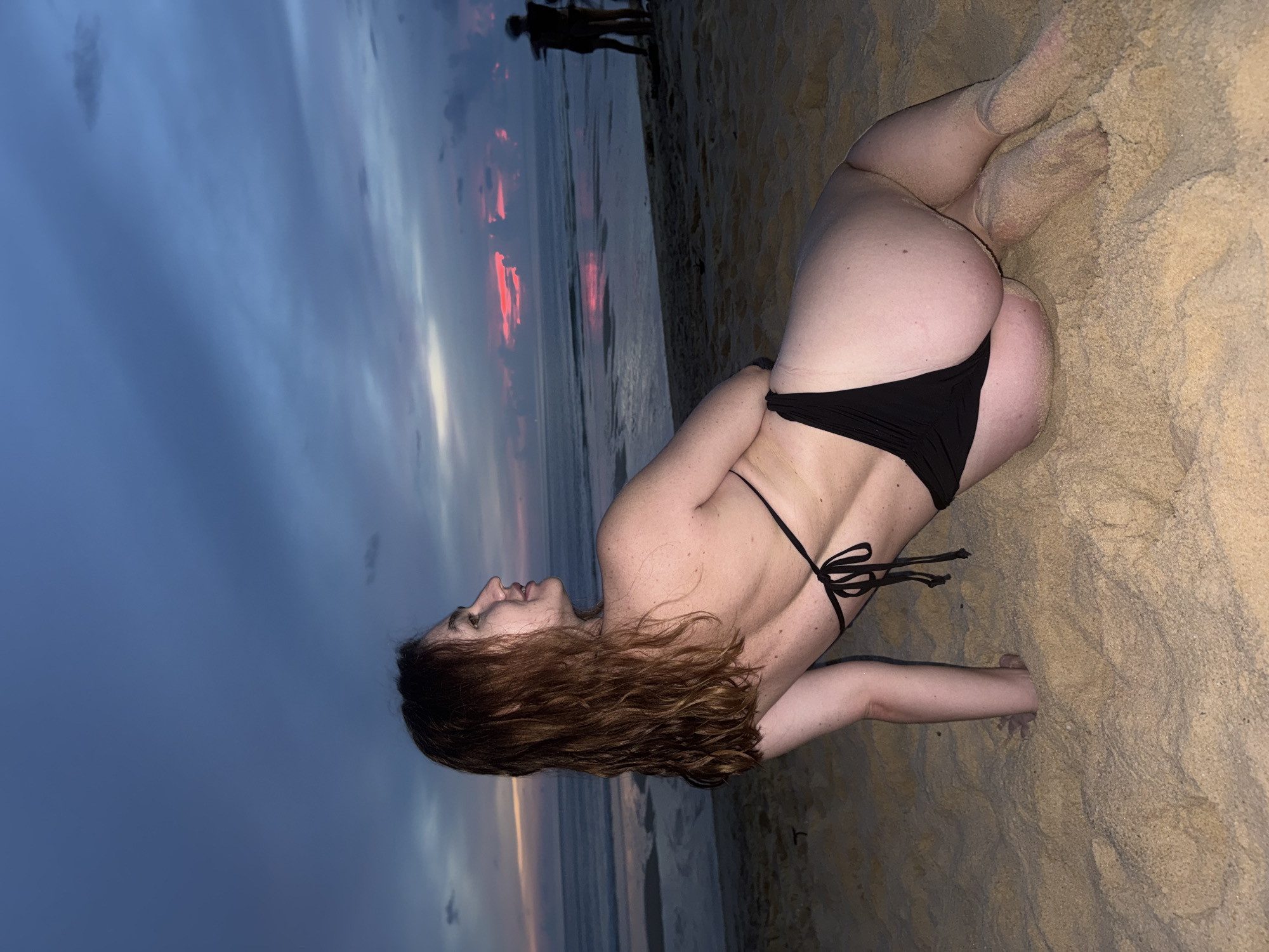 A woman in a black bikini is sitting on a sandy beach in Choeng Thale, Thailand, looking out at the ocean. The sky is a mix of deep blue and pink hues, indicating either sunrise or sunset. The ocean has gentle waves, and the sand is light brown. In the background, two figures can be seen standing near the water's edge. The woman is facing away from the camera, with her back towards the viewer, her head turned to her left.