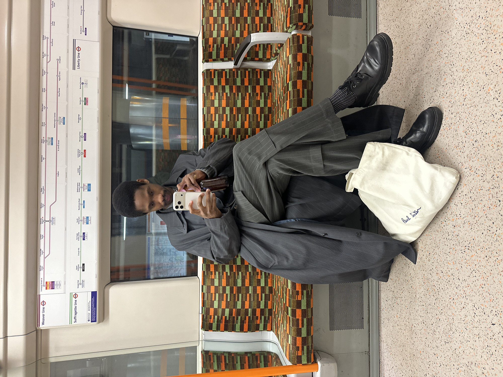 A man wearing a grey pinstripe suit and black shoes sits with his legs crossed in a London Underground train carriage. He is holding a smartphone. The seat has a patterned fabric. Next to him is a white Paul Smith tote bag. A London Underground map is on the wall above the window. The map indicates the train is on the 'Weaver Line' and the 'Suffragette Line'. Place names such as Liverpool Street, Hackney, Crouch Hill, Southbury, Walthamstow Central, St. James Street, Wood Street, Highams Park, Chingford, Liberty Line, Barking, and Emerson Park are visible.