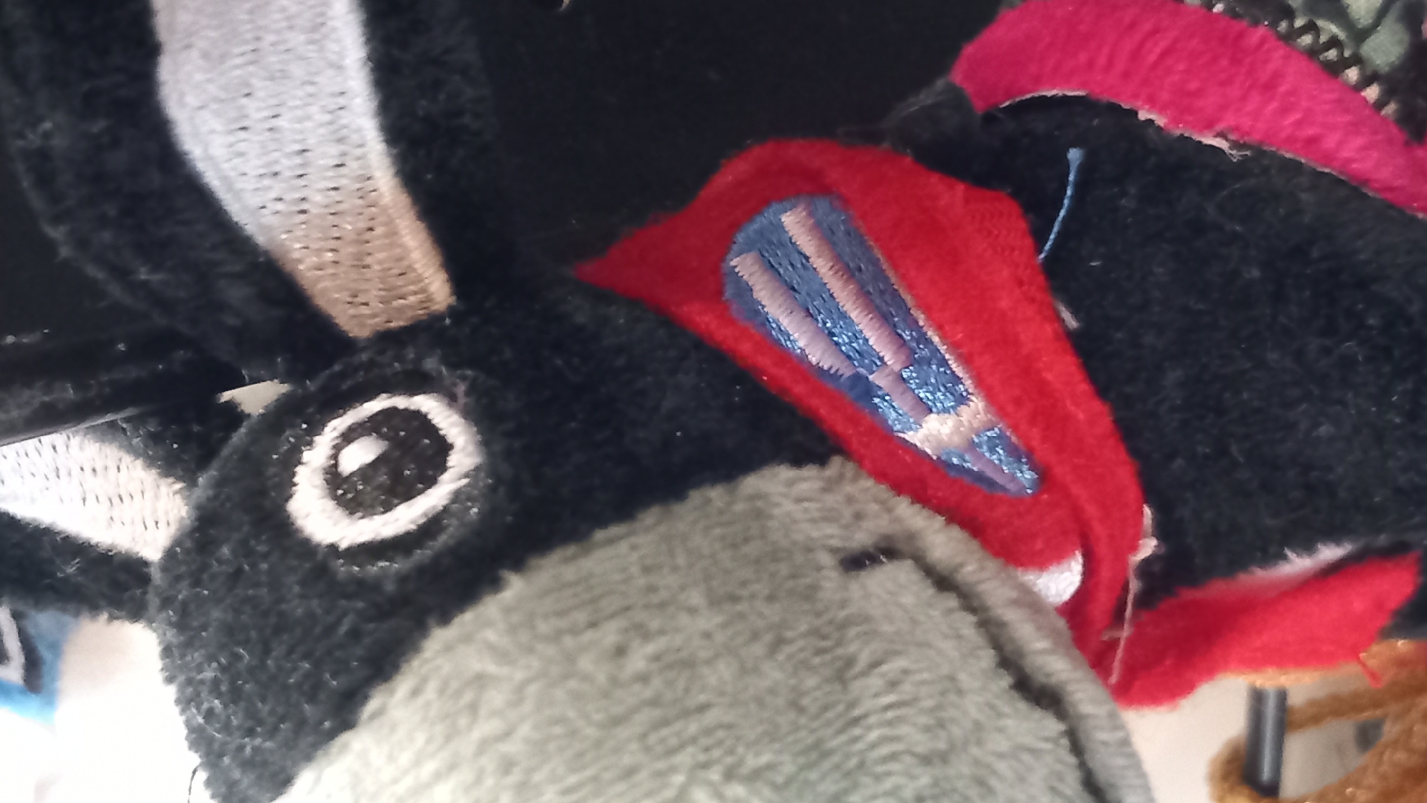 The image shows a close-up of a stuffed toy, likely a donkey, with black fur and a gray snout. The toy has a white and black embroidered eye, and a red collar with blue and pink embroidered details. A thin, light-colored band with a woven texture is visible across the toy's head. The background is dark and out of focus. There is no information in the image to suggest its location in the city of Kos, Greece.