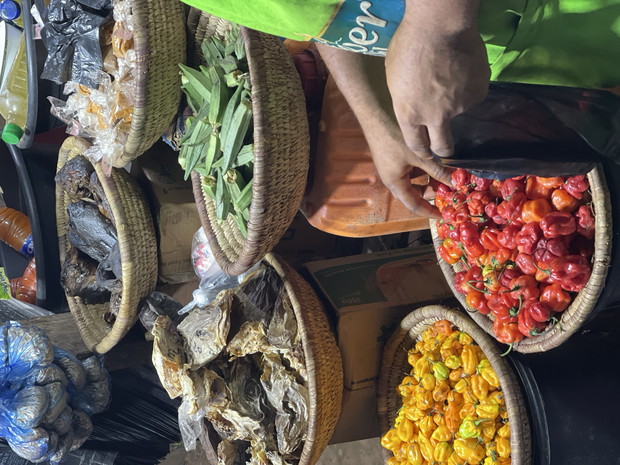 This overhead shot captures a vibrant and busy market stall, likely in Abuja, Nigeria. The scene is dominated by multiple hand-woven wicker baskets filled with various foodstuffs and ingredients.
In the upper-left, a basket holds numerous small plastic bags containing an orange-colored powder, possibly spices. Below this, another basket contains dried fish or seafood, characterized by a dark, shriveled appearance. Further down and to the left, blue plastic bags tightly packed with an unidentified product, possibly "POLY" as indicated by visible text, are stacked. Adjacent to these, another basket also holds dried fish, this time appearing lighter in color and flakier.
To the right, a large basket is filled with fresh green okra pods. Below this, two baskets are brimming with colorful peppers: the upper one with red and orange Scotch bonnets, and the lower one with bright yellow and orange varieties. A person, wearing a green shirt with visible text "er" (possibly part of "Super" or "Power") and a blue patterned sleeve, is seen in the upper right, reaching into the basket of red peppers, suggesting a transaction or preparation for sale.
Notable details include a yellow plastic bottle with a green cap, likely containing oil, and an orange bottle in the background on the left. Various cardboard boxes are visible, serving as display or storage, with legible text such as "350g Pouch Instant Milk Powder" and "Campina" on one box, and "Good, Good life" and "UP THIS SIDE OF" on others. The blue-wrapped item near the bottom left has "Fresh Garlic" written on a label. The lighting suggests an indoor market or an evening setting.