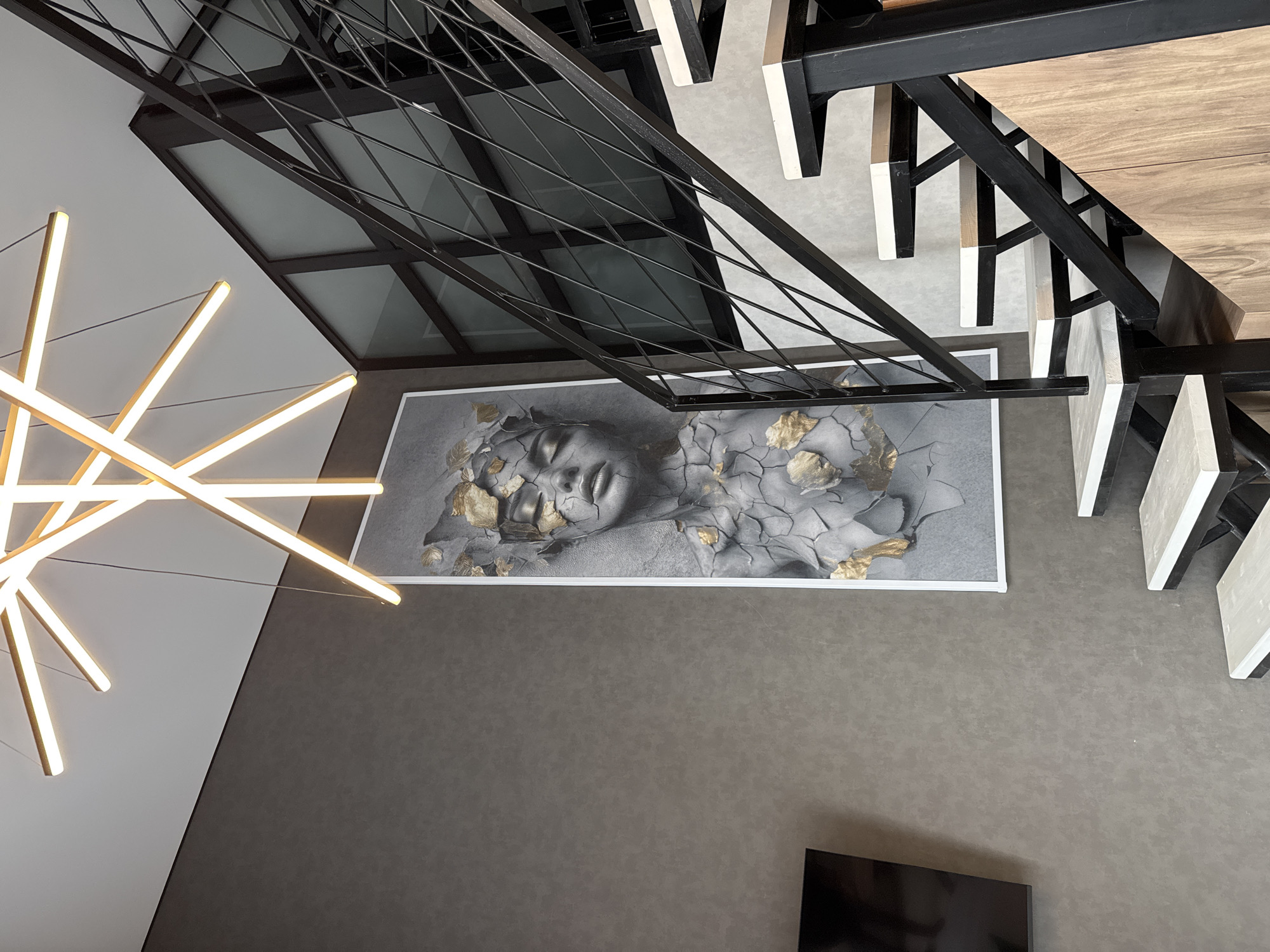 This image presents a modern interior space, viewed from an elevated perspective looking downwards. The primary subjects include a staircase, a unique light fixture, and a large piece of wall art.
On the left, a large, abstract chandelier with multiple glowing, golden-toned bars extends across a light grey wall section. This wall transitions into a darker, textured grey-brown surface that dominates the lower and central areas of the image.
A prominent black metal staircase with wooden treads ascends from the lower right towards the upper right. Its railing features a distinctive criss-cross geometric pattern. Below the staircase and centrally positioned on the dark grey-brown wall is a large framed artwork. This artwork depicts a face, possibly a woman's, with closed eyes, appearing to break apart, revealing gold leaf accents within the cracks and on the surface.
The floor or lower level visible beneath the staircase is a light grey, possibly concrete. A corner of a black television screen is discernible in the bottom right. The overall aesthetic is contemporary and sophisticated, utilizing a muted color palette of greys, black, natural wood, and striking gold highlights from both the light fixture and the artwork. No people or visible text are present in the image.