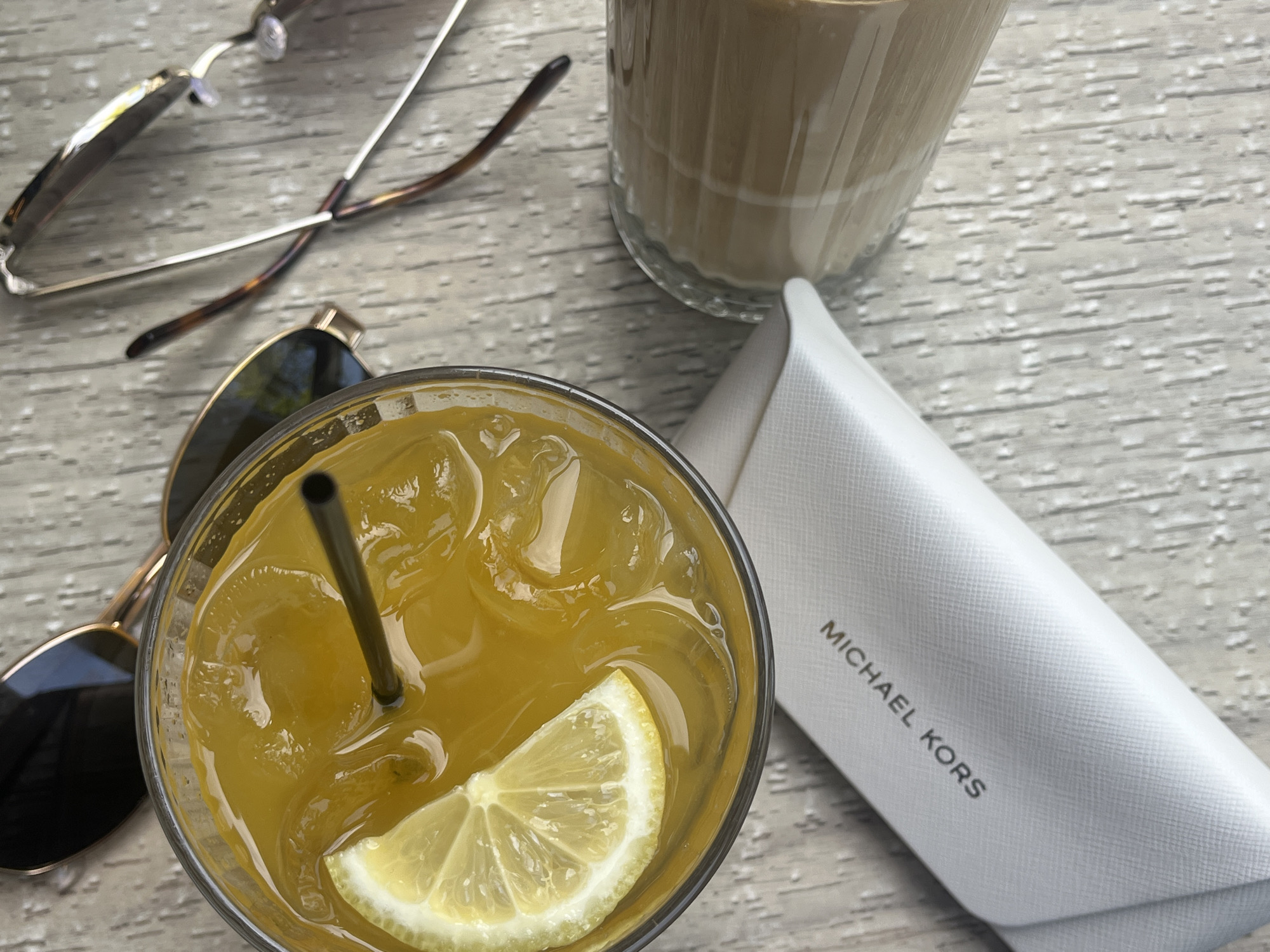 The image captures a high-angle, close-up view of a casual table setting, likely outdoors or in a bright indoor space. The main surface is a light-colored, textured wooden table.
In the foreground, a clear glass holds a vibrant yellow-orange iced beverage, featuring a dark metal straw and a fresh lemon slice. To its left, a pair of sunglasses with dark, rectangular lenses and gold-colored frames rests on the table.
Towards the top right, another glass contains a layered beverage, appearing to be an iced coffee or latte with distinct beige and white layers. Partially visible in the top left is a second pair of sunglasses, characterized by thin silver frames and brown tortoise-shell patterned arms.
A prominent white, rectangular case or pouch, displaying a subtle textured pattern, is situated in the mid-right. Clearly visible on its surface in black capital letters is the text "MICHAEL KORS."
The scene suggests a relaxed daytime activity, possibly enjoying drinks at a cafe or restaurant, with the presence of sunglasses indicating bright or sunny weather. While the contextual location is Plovdiv, Bulgaria, no specific visual elements within the image itself confirm this setting. The overall atmosphere is one of leisure and enjoyment.