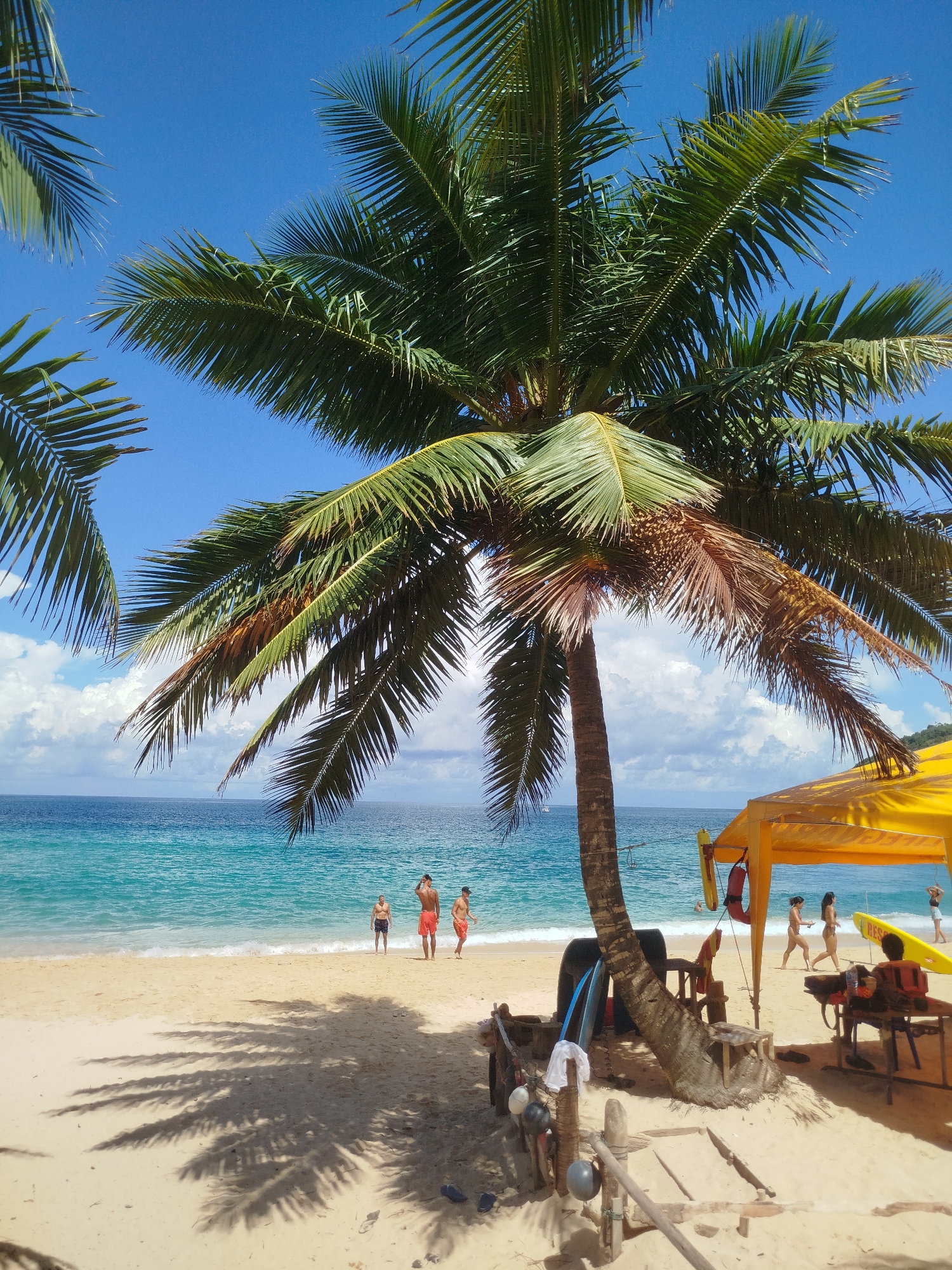 This image depicts a sunny day at a beach in Rawai, Thailand. A prominent palm tree with green and brown fronds dominates the foreground, casting long shadows on the sand. To the right, a yellow canopy shelters a lifeguard station with equipment like a life preserver and a rescue buoy. Several people are visible: three men stand near the water's edge in the mid-ground, while others, including women in swimwear, are seen walking along the beach near the lifeguard station. The ocean is a vibrant turquoise, with small waves breaking on the shore. The sky is a clear blue with scattered white clouds. The beach is sandy and appears to be a popular spot for recreation.