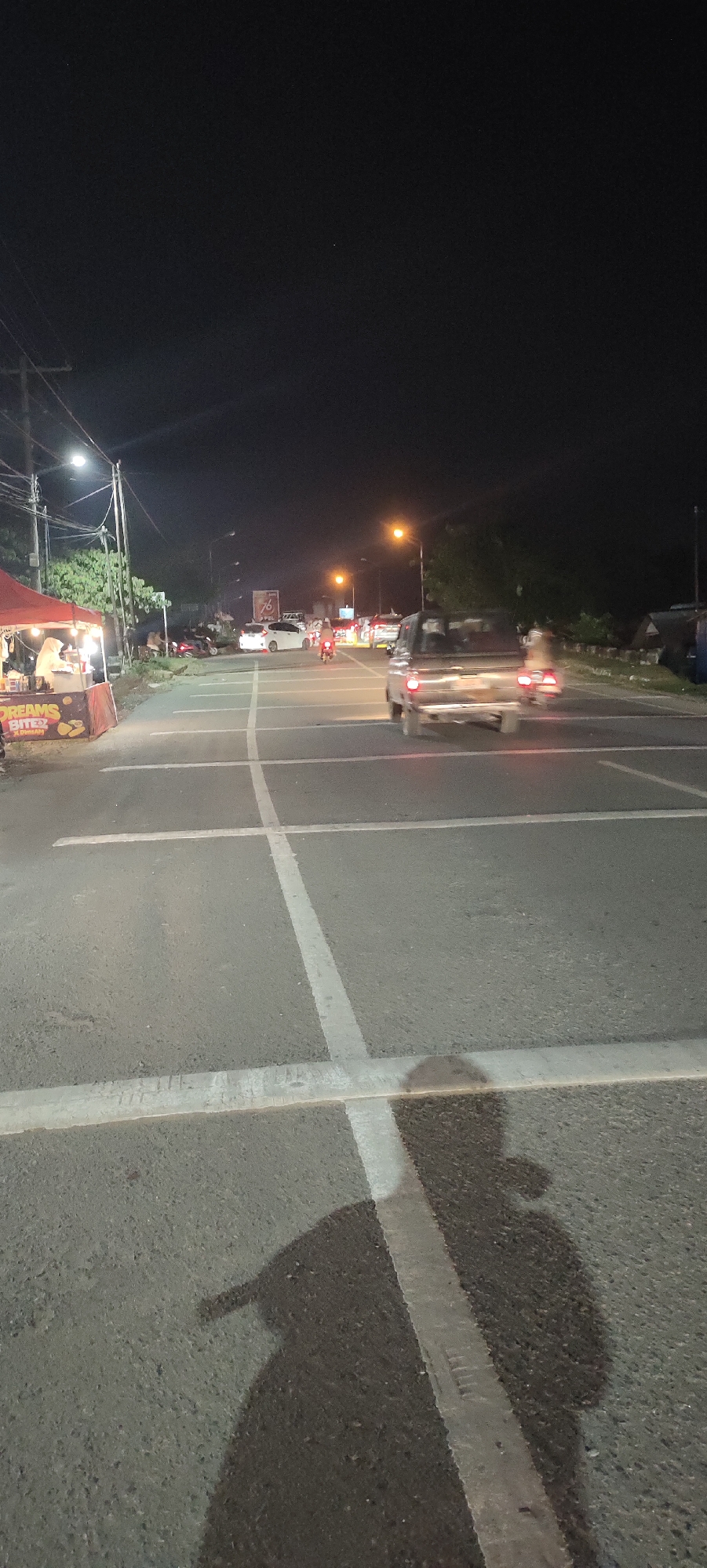 At night in Lhokseumawe, Indonesia, a road intersection with white painted lane markings is illuminated by streetlights. A food stall sits to the left of the road with a red awning. Vehicles, including cars and motorcycles, are moving across the intersection. A shadow of a person holding a camera stretches across the lower portion of the road, indicating the perspective of the photographer.