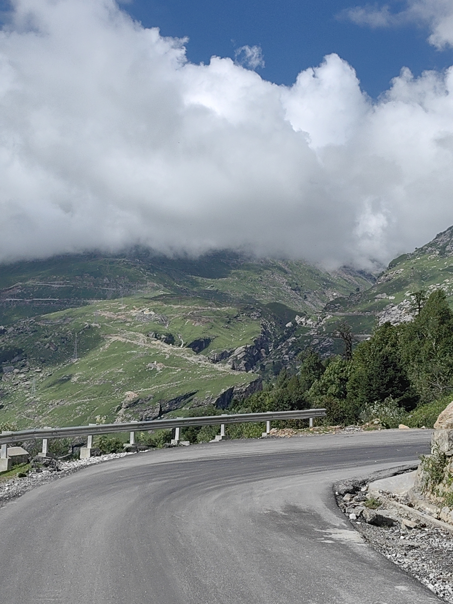 A winding paved road curves through a mountainous landscape in Kothi, India. The road is bordered by a metal guardrail supported by concrete pillars. The mountainside is covered in green grass and scattered rocks, with a winding road visible higher up the slope. Dark green trees are present on the right side of the image. The sky is blue with fluffy white clouds.
