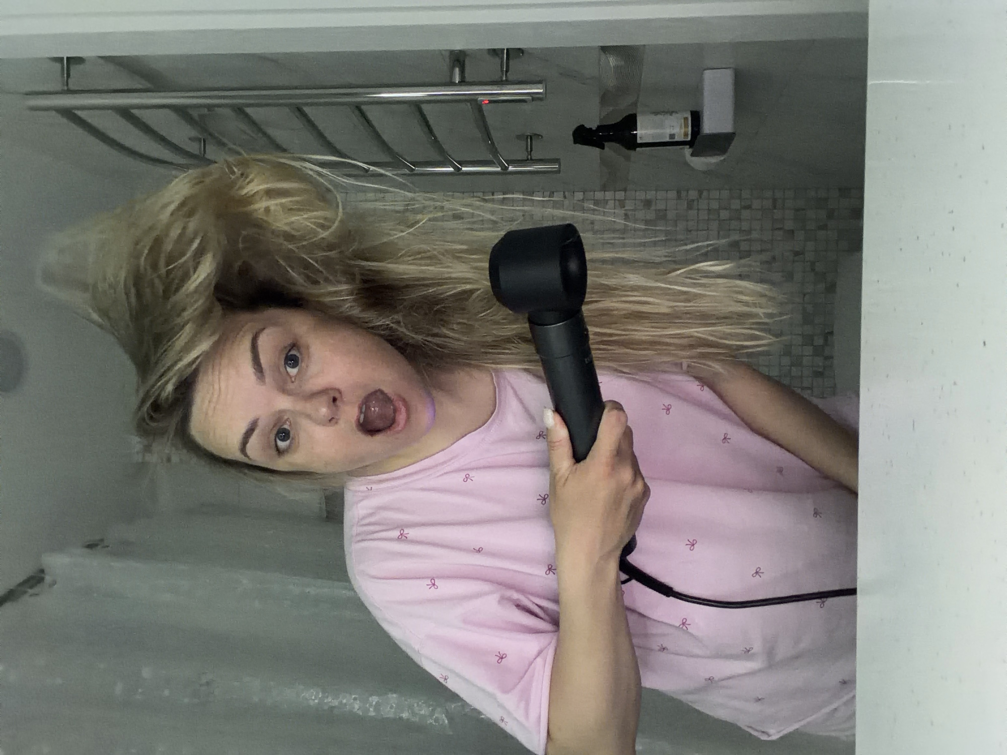 A woman is holding a black hair dryer in a bathroom. The woman has light blonde hair and is wearing a light pink t-shirt with a small bow pattern. She is looking at the camera with her mouth open. Behind her is a metal towel rack, a bottle, and a tiled wall. The room appears to be a bathroom due to the presence of a shower curtain on the left side of the frame. The location is not Votkinsk, Russia. The location cannot be determined from the given information.