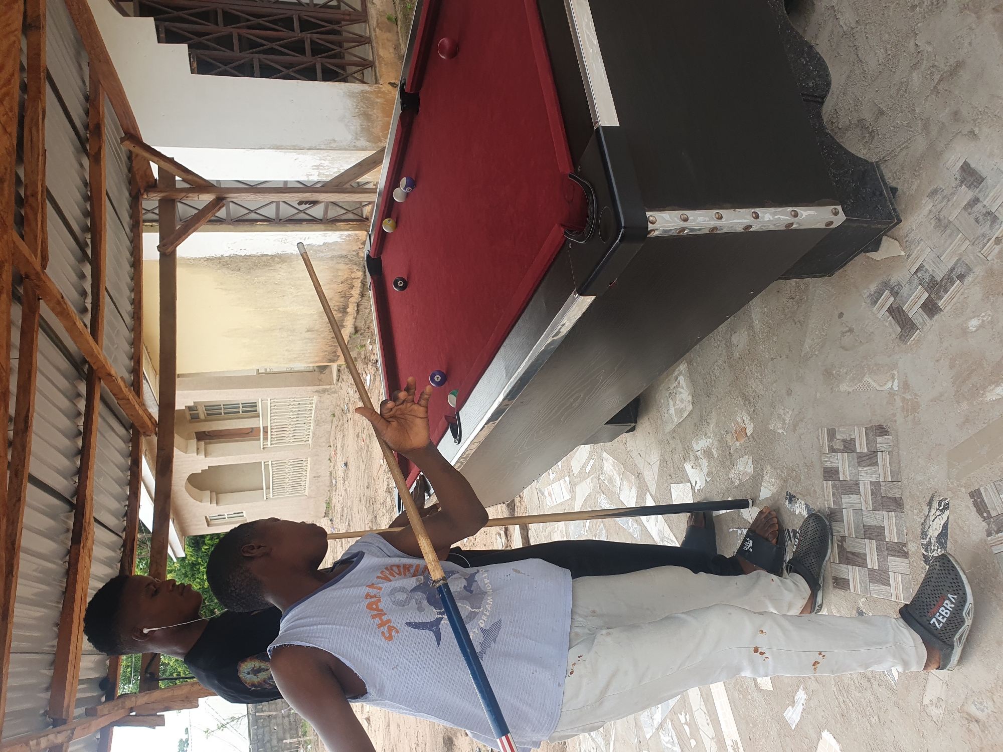 In Lokoja, Nigeria, two young men are playing pool outdoors under a wooden, corrugated metal roof. The man in the foreground, wearing a white tank top with a graphic print and light-colored pants, is in the process of striking a billiard ball with a pool cue. His right arm is extended, holding the cue horizontally, and his body is angled towards the pool table. A second man, wearing a black t-shirt and black pants, stands behind him, observing the game and also holding a pool cue vertically. The pool table is dark with a red felt surface, and several billiard balls are scattered across it. The ground is paved with patterned tiles in some areas and dirt in others. In the background, a light-colored building with arched windows and white railings is visible. The overall setting appears to be an informal, outdoor recreational area.