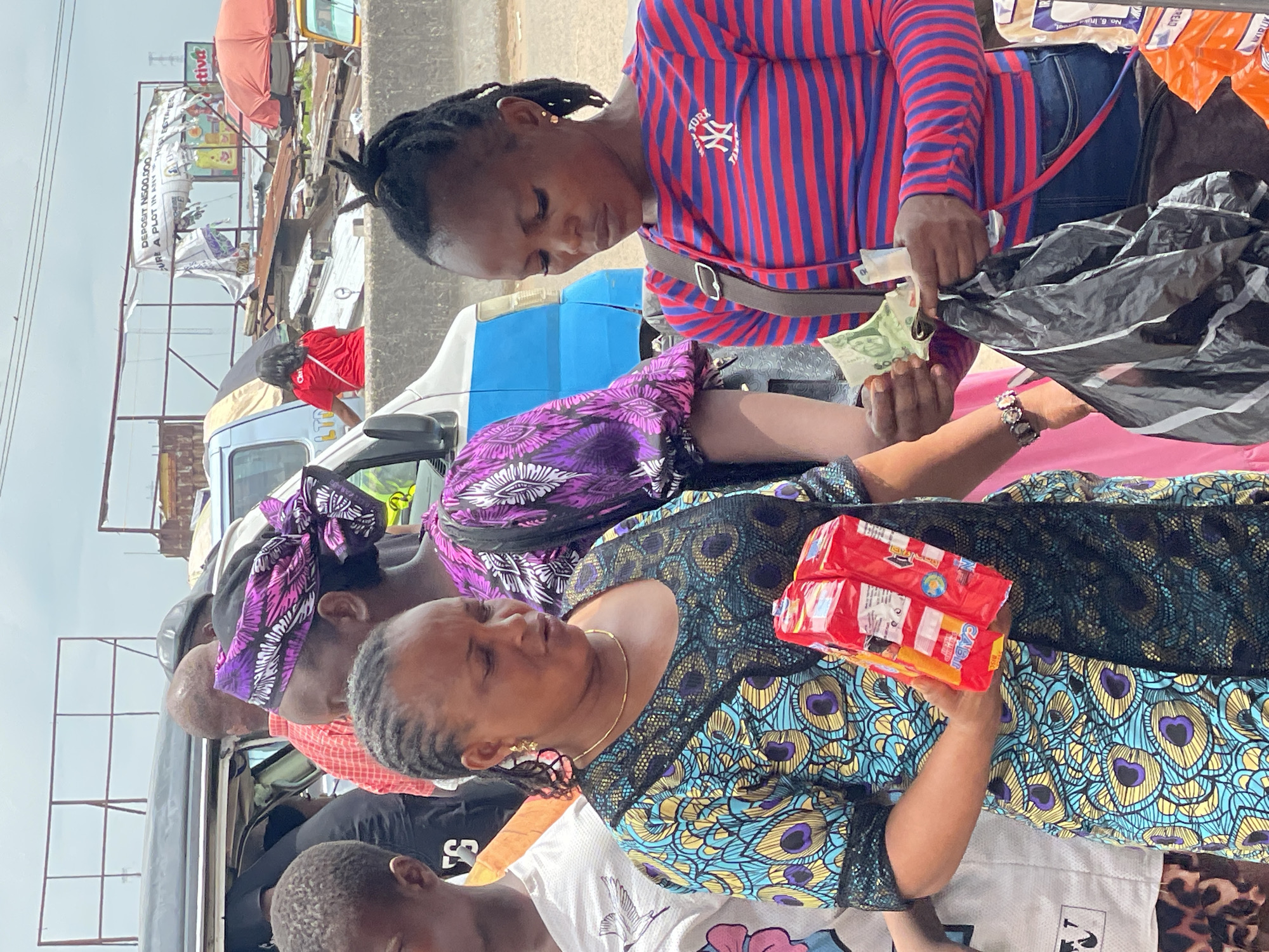 The image shows a street scene in Onitsha, Nigeria, where a woman wearing a brightly patterned blue and gold dress is making a purchase from another woman dressed in a red and blue striped shirt. The woman in the patterned dress holds two red packages labeled "CABI" as she receives change from the other woman, who is counting Nigerian naira notes from a black plastic bag. A van with blue accents is visible in the background, along with other people. There's also a billboard in the background advertising land plots for sale, mentioning a deposit amount and real estate-related text. The overall environment suggests a busy market or street vending area.