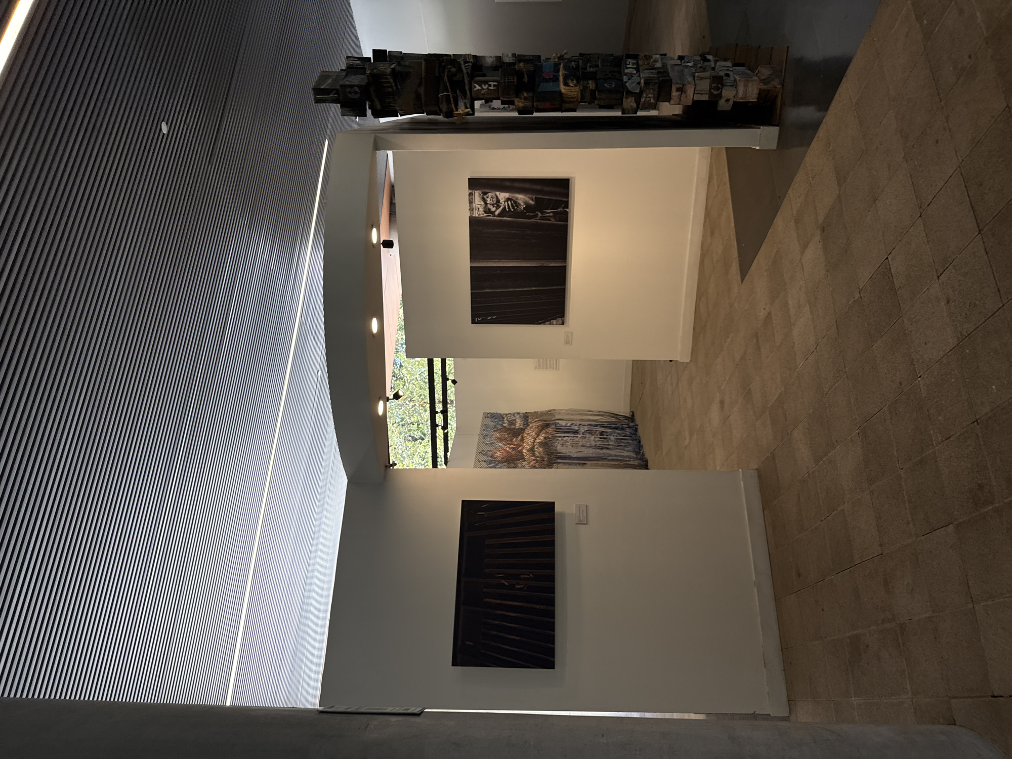 The image captures the interior of a modern art gallery or exhibition space, characterized by striking architecture and a curated display of artworks. The dominant feature on the left is a high, angled wall and ceiling clad in horizontal wooden slats or panels, creating a textured surface. Recessed strip lighting runs along the angles, illuminating the space, complemented by a few circular spotlights.
The gallery space features white walls presenting three distinct artworks. At the top, a horizontal sculpture or assemblage crafted from numerous irregular wooden blocks or printer's type, some with faintly visible letters and symbols such as "HAY," is mounted above a white partition. Below it, a framed black and white photograph depicts what appears to be a person or figure in a somber setting, with a small, unreadable inscription in the bottom left corner, possibly "10-20." Further down, a dark, rectangular artwork, possibly a painting or mixed media piece with horizontal elements, hangs on the wall. Another large, abstract painting or tapestry, primarily in blues and browns, is visible on an upper level through an opening in the wall, with green foliage from outside visible beyond it.
The floor is a light brown, tiled surface laid in a grid pattern. The overall atmosphere is calm and contemplative, typical of an art exhibition. Natural light from an unseen window or opening on the right side indicates it is daytime. Small white labels are present next to the artworks, but their text is not discernible. The setting suggests a contemporary cultural institution, likely in Mexico City as per the provided context.